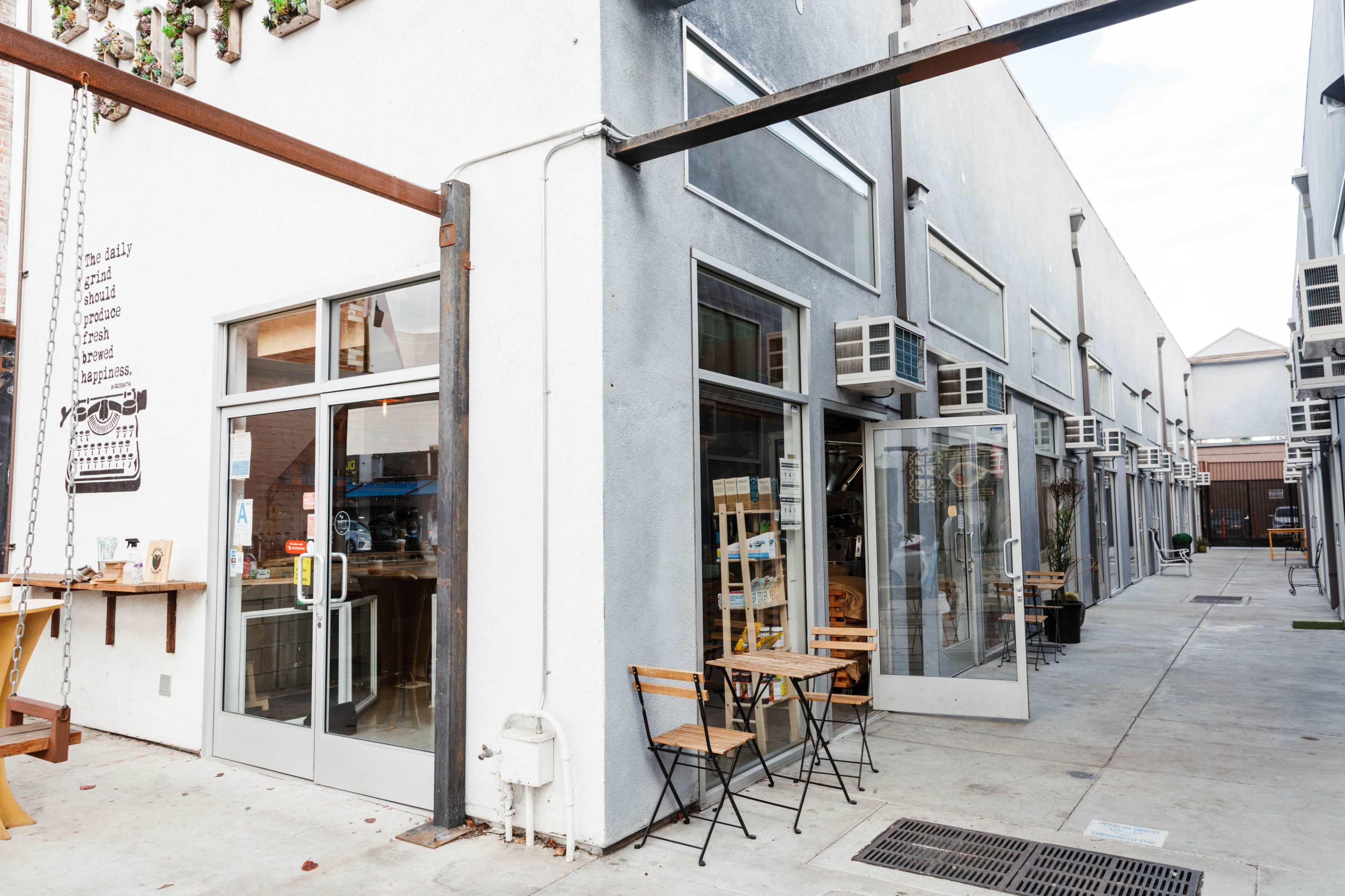 The image shows a modern retail space with large glass windows, outdoor seating, and a concrete walkway lined with additional shops.