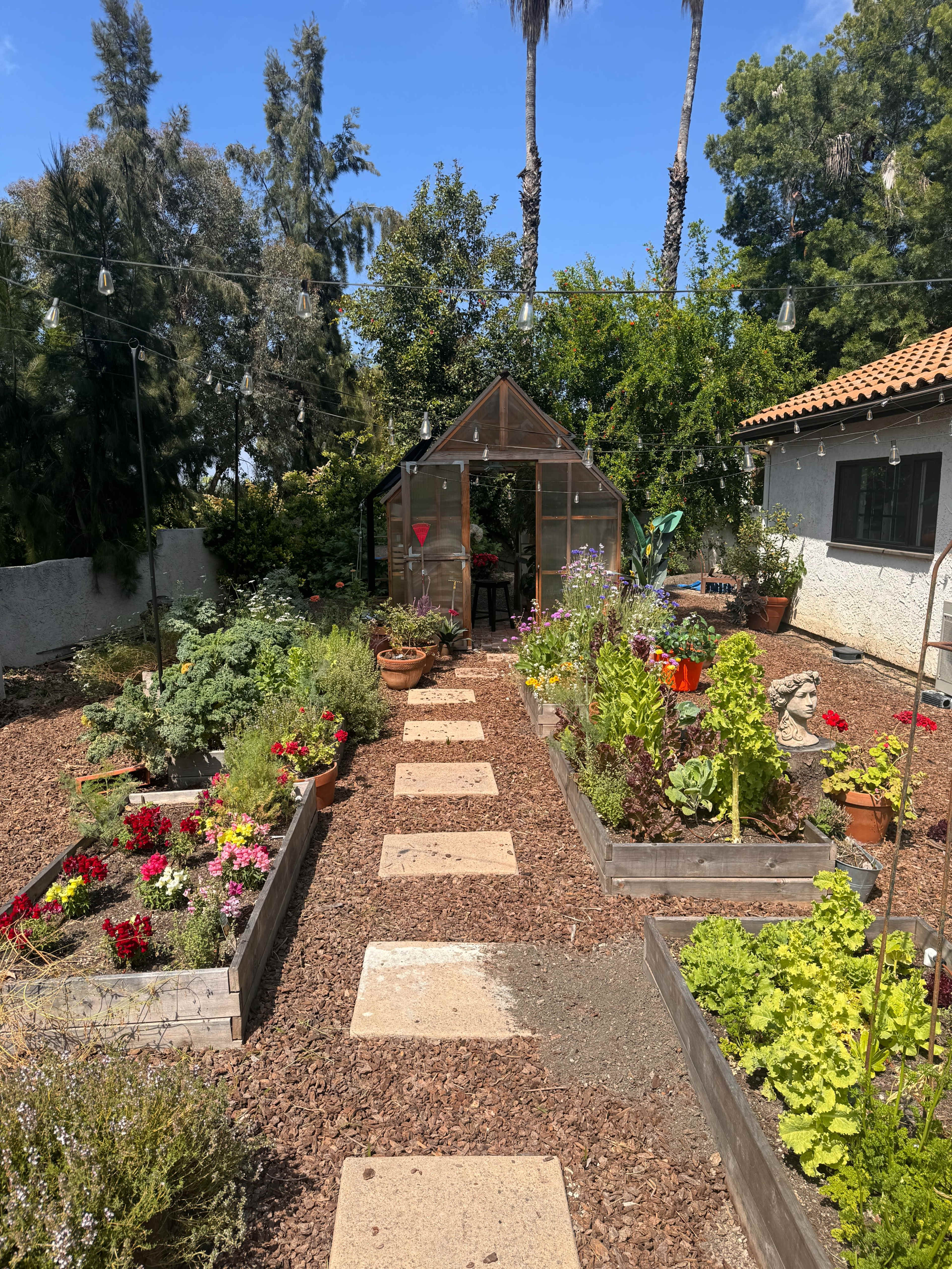 A garden path lined with stepping stones leading to a greenhouse, surrounded by flowerbeds and vegetable patches under a clear sky.