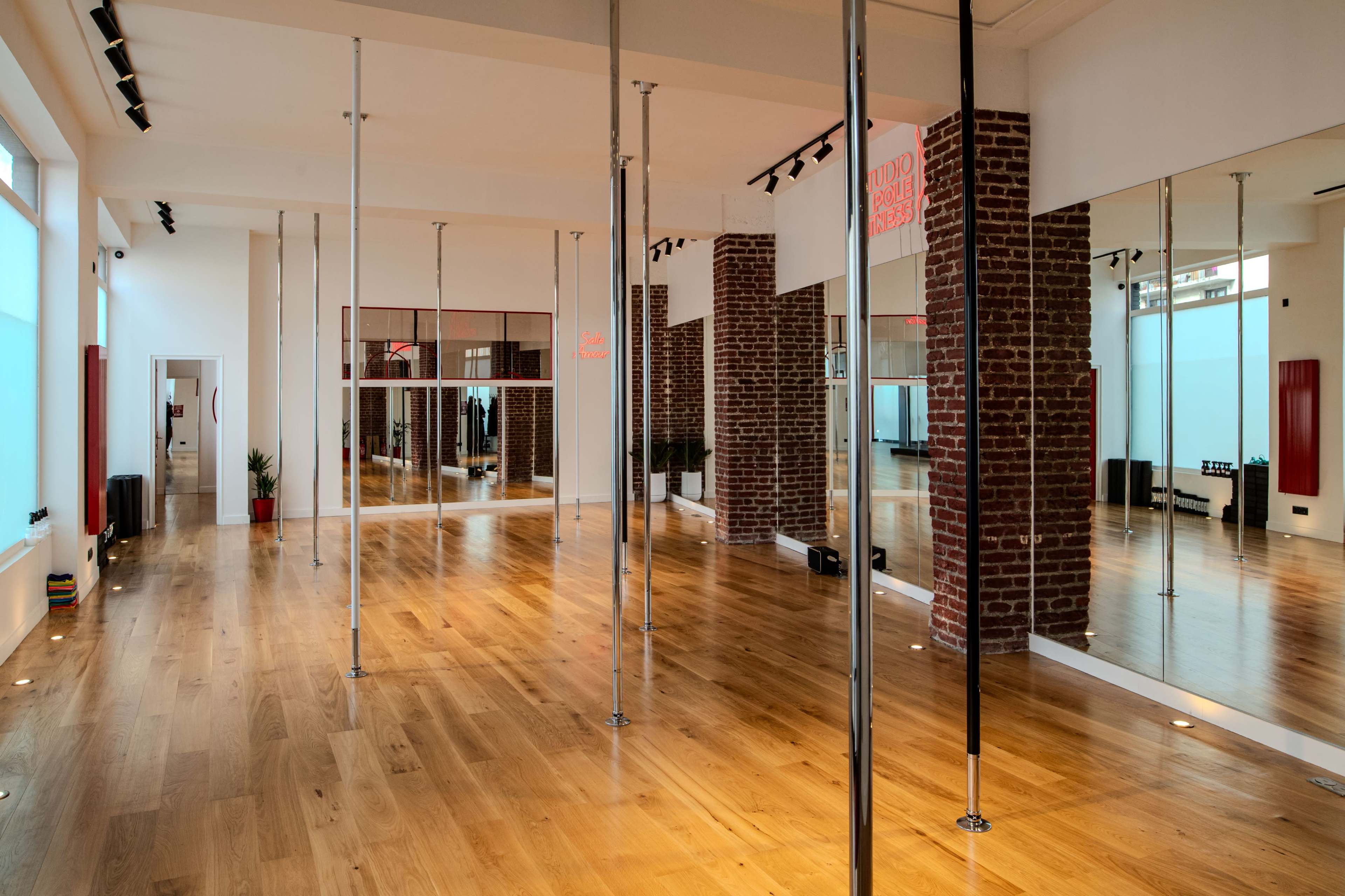 The image displays a spacious dance studio featuring several vertical poles, large mirrors, and wooden flooring, with a brick wall on one side.