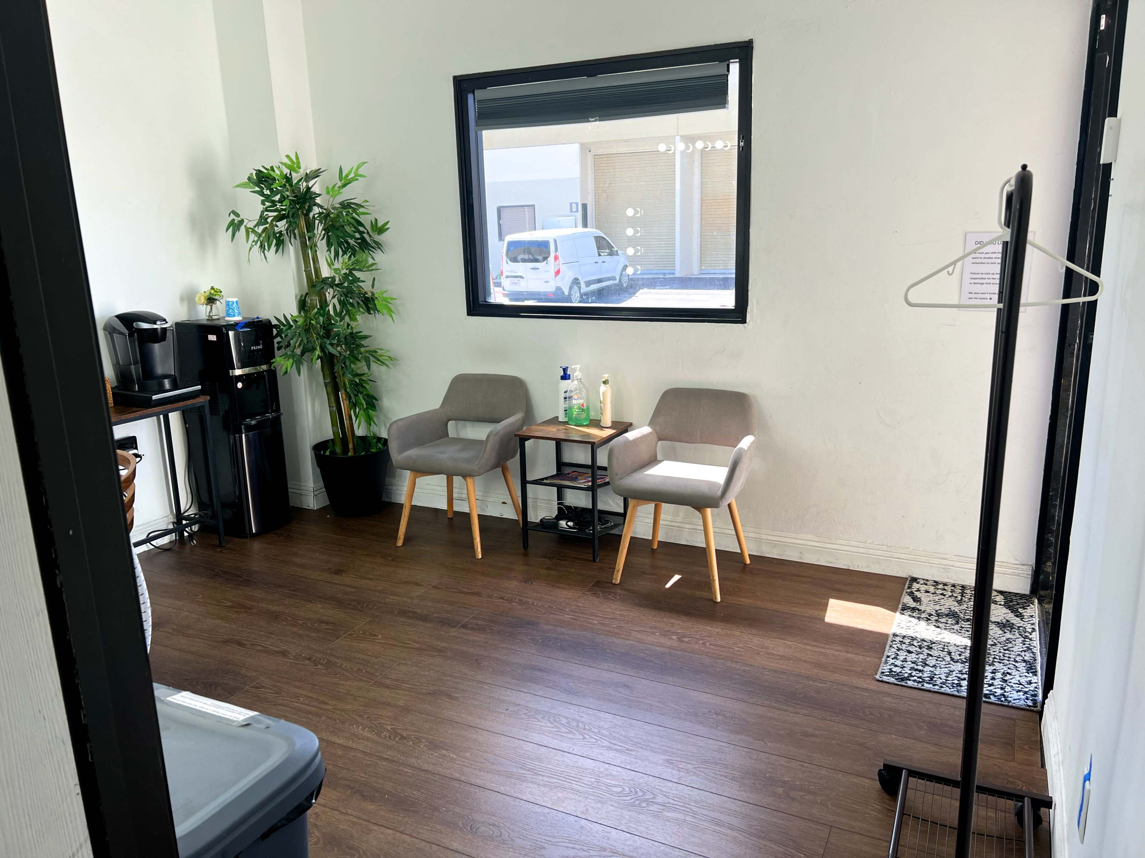 The image shows a small room with two gray chairs, a side table, a potted plant, and a coffee station along one wall.