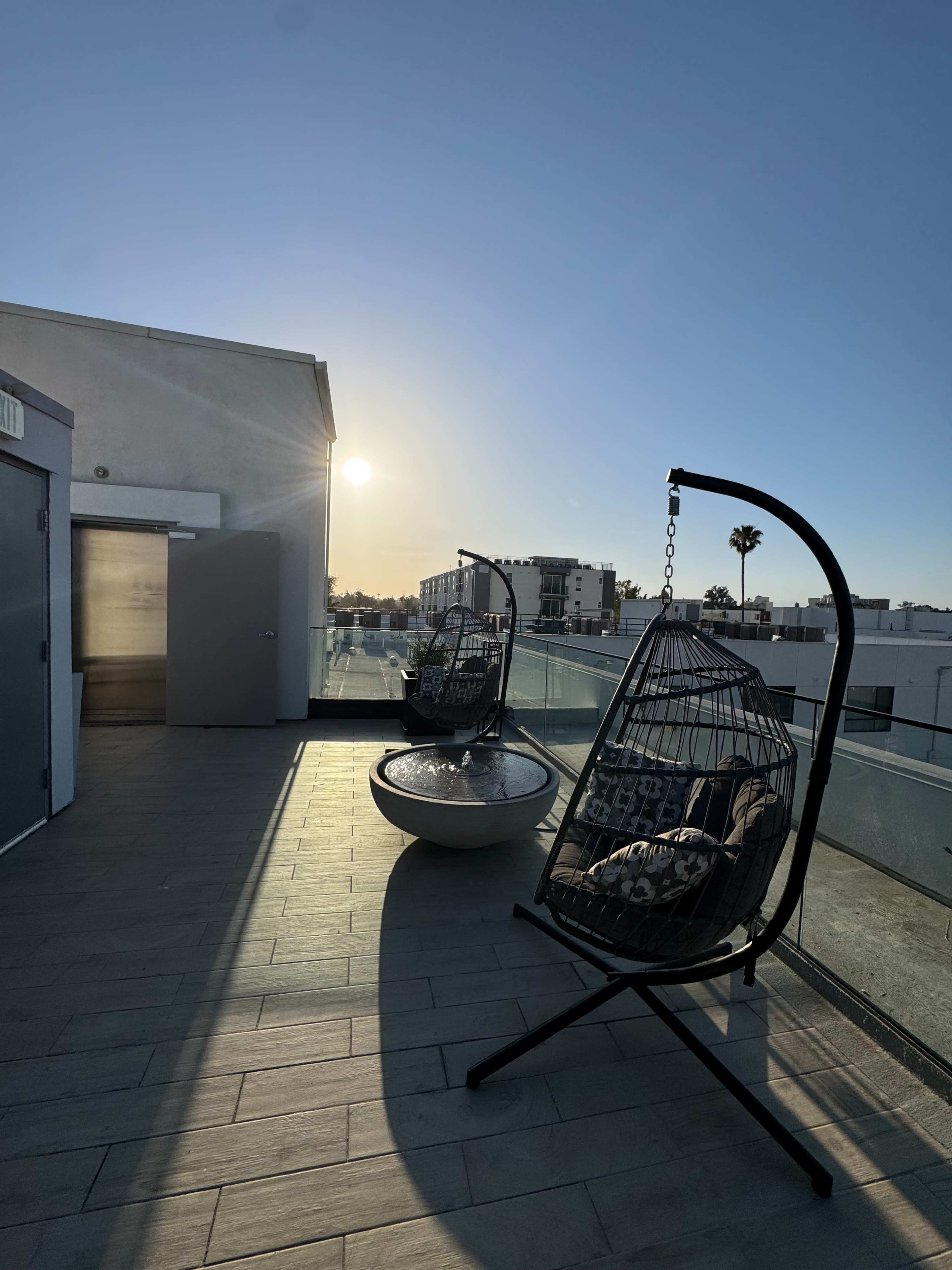 Two hanging chairs are positioned on a rooftop terrace, with a view of the setting sun and nearby buildings.