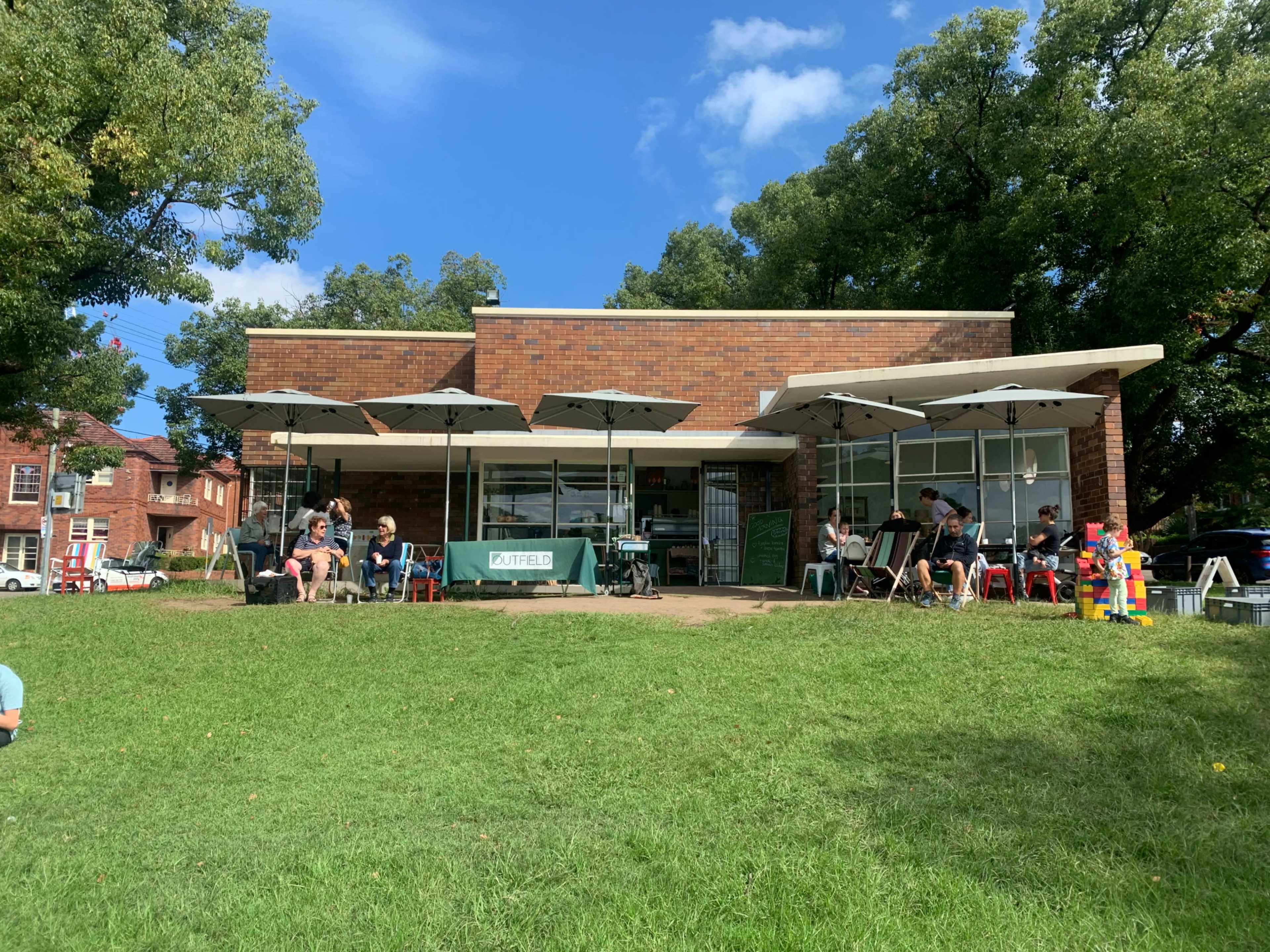 A brick cafe with outdoor seating shaded by umbrellas is situated on a grassy area, surrounded by trees.