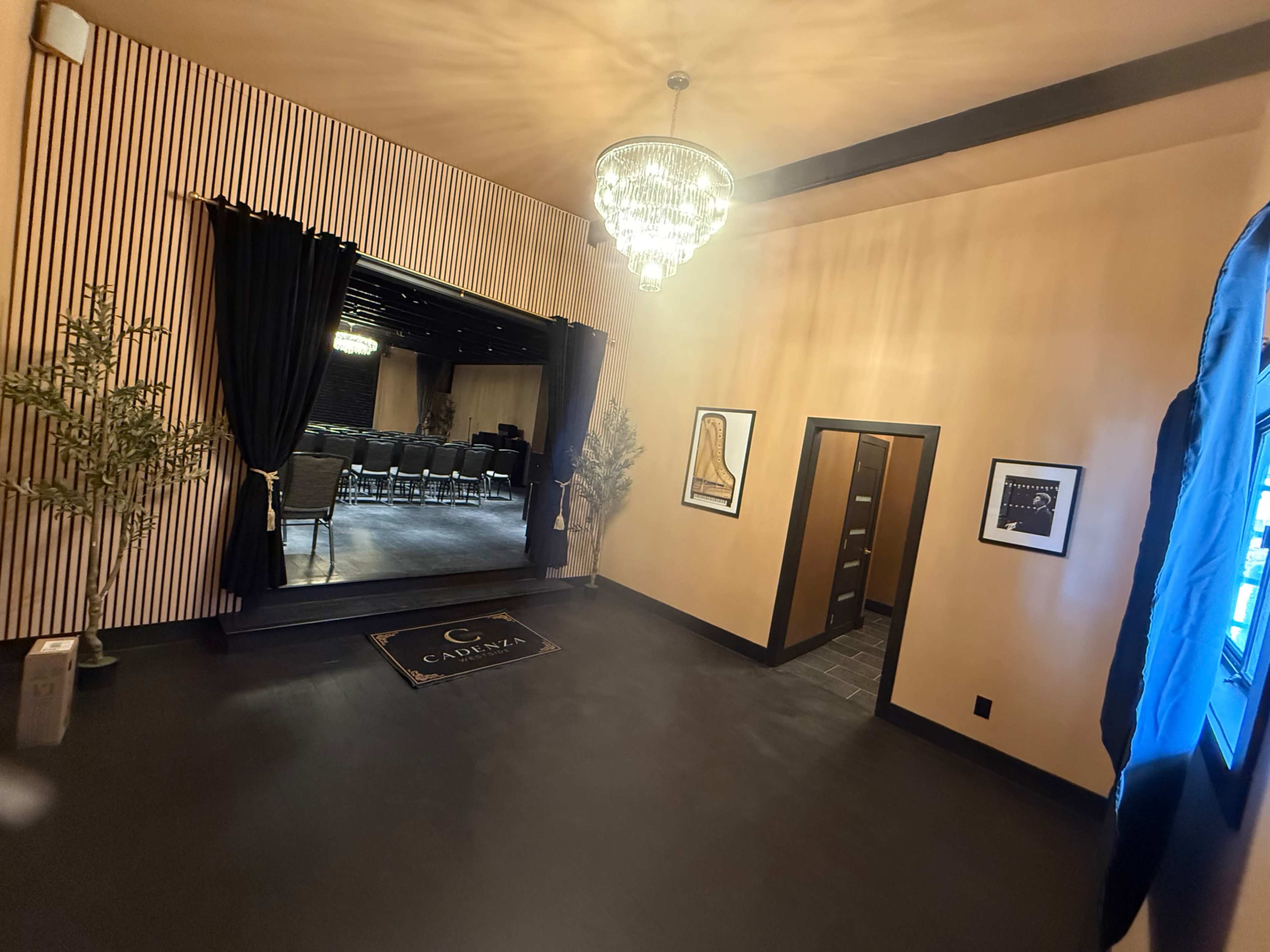 The image shows a spacious event room with a black floor, beige walls, a chandelier, and a stage area draped in black curtains, featuring a seating arrangement in the background.