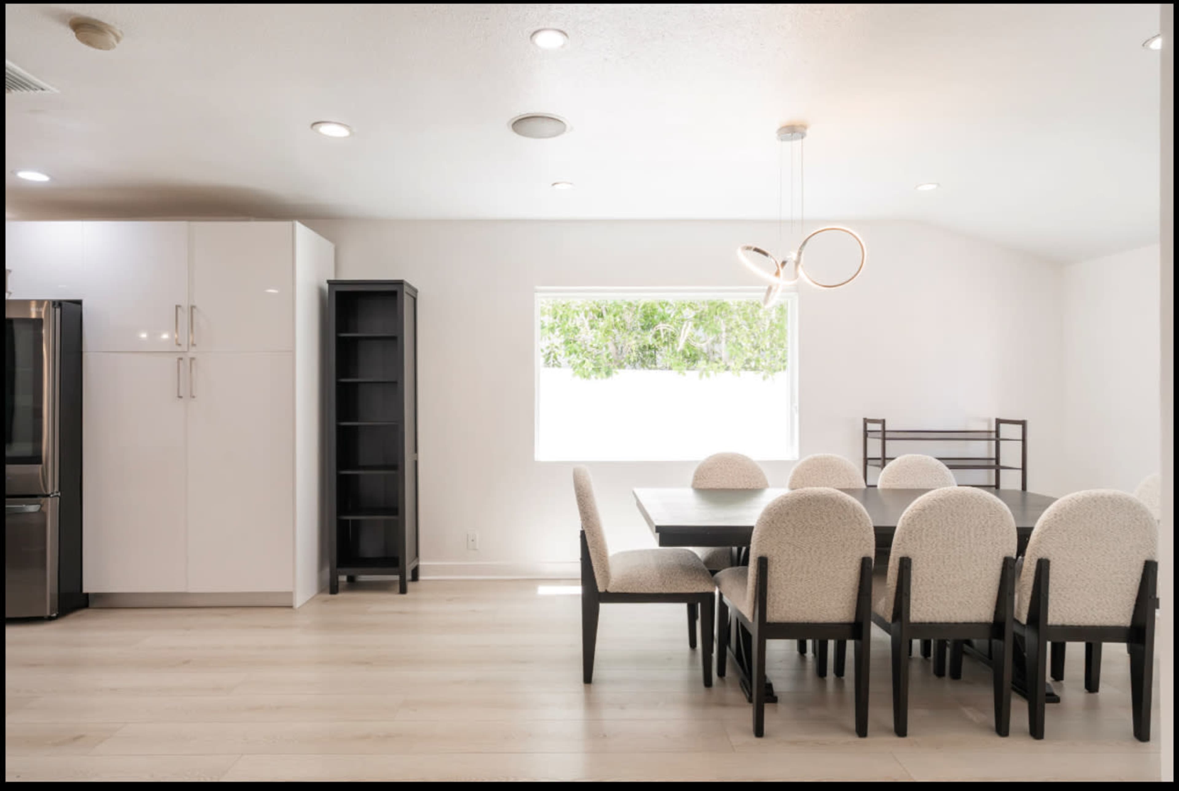 A dining room features a large table surrounded by upholstered chairs, with a window providing natural light and sleek, modern furniture.