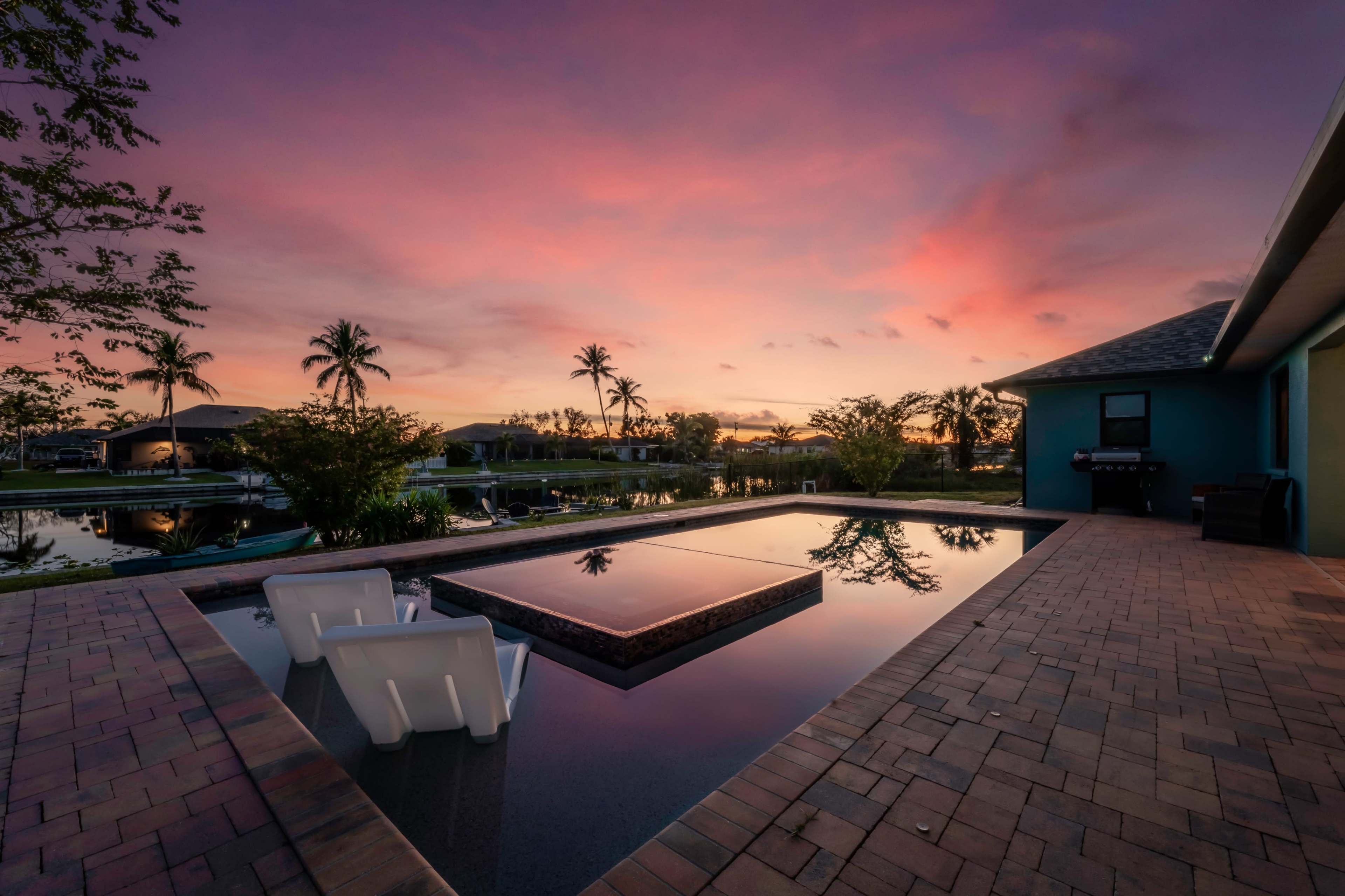 A serene pool area with white chairs, surrounded by lush vegetation and a colorful sunset reflecting on the water's surface.