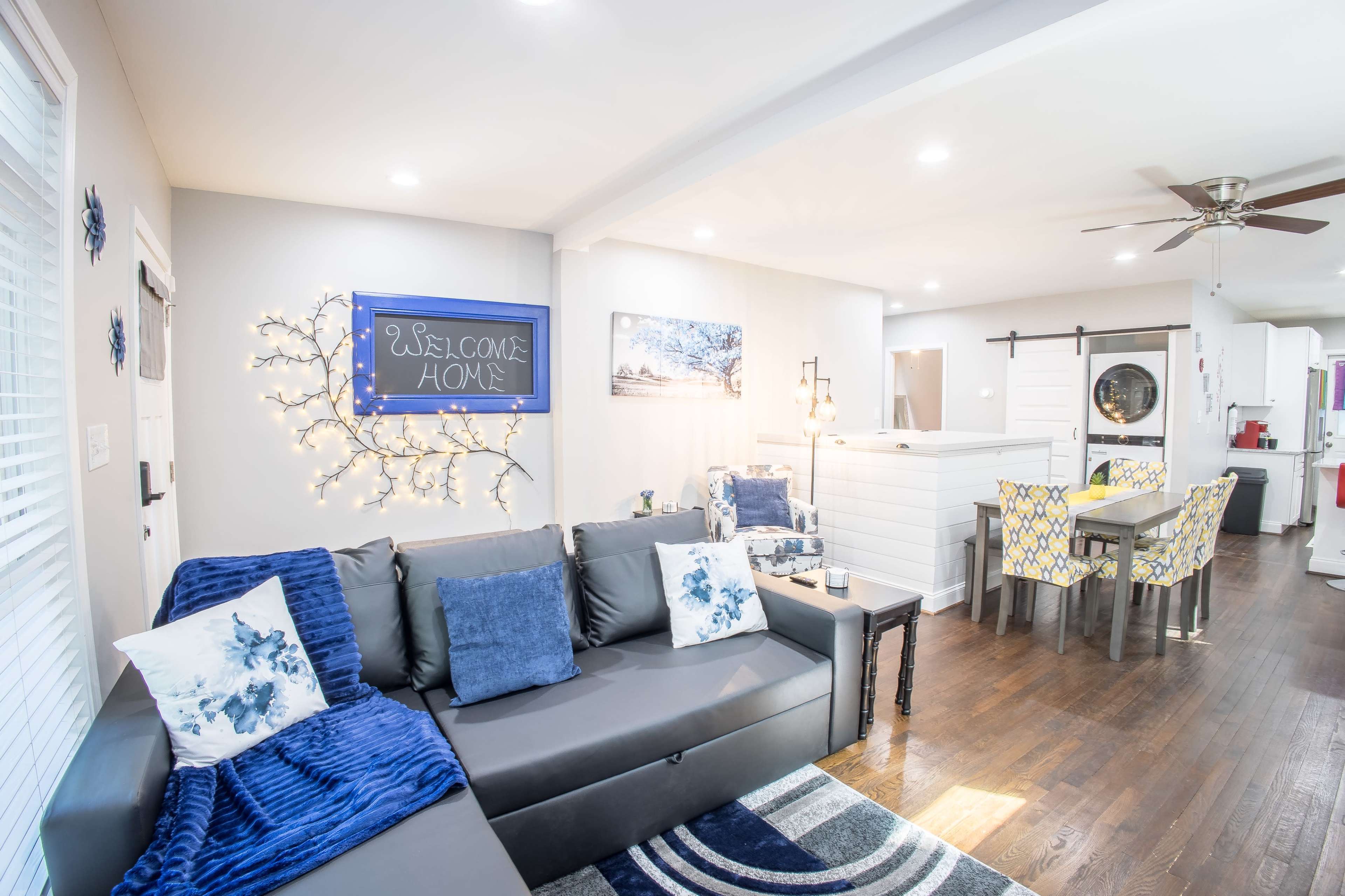 A modern living room with a gray sofa, a welcome sign on the wall, and a dining area in the background.