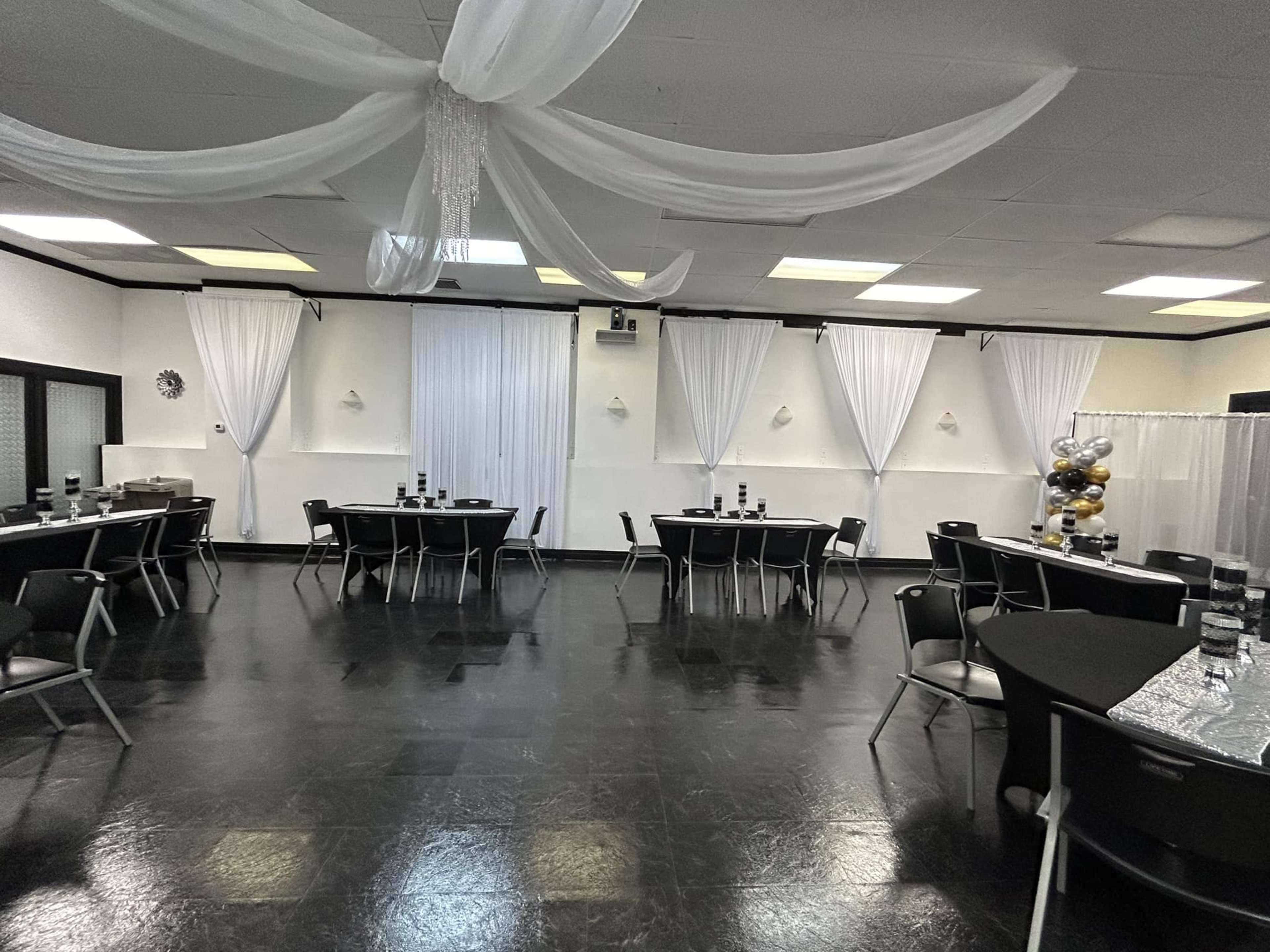 A spacious event room featuring black tables and chairs arranged around an elegantly decorated floor with white drapes hanging from the ceiling.