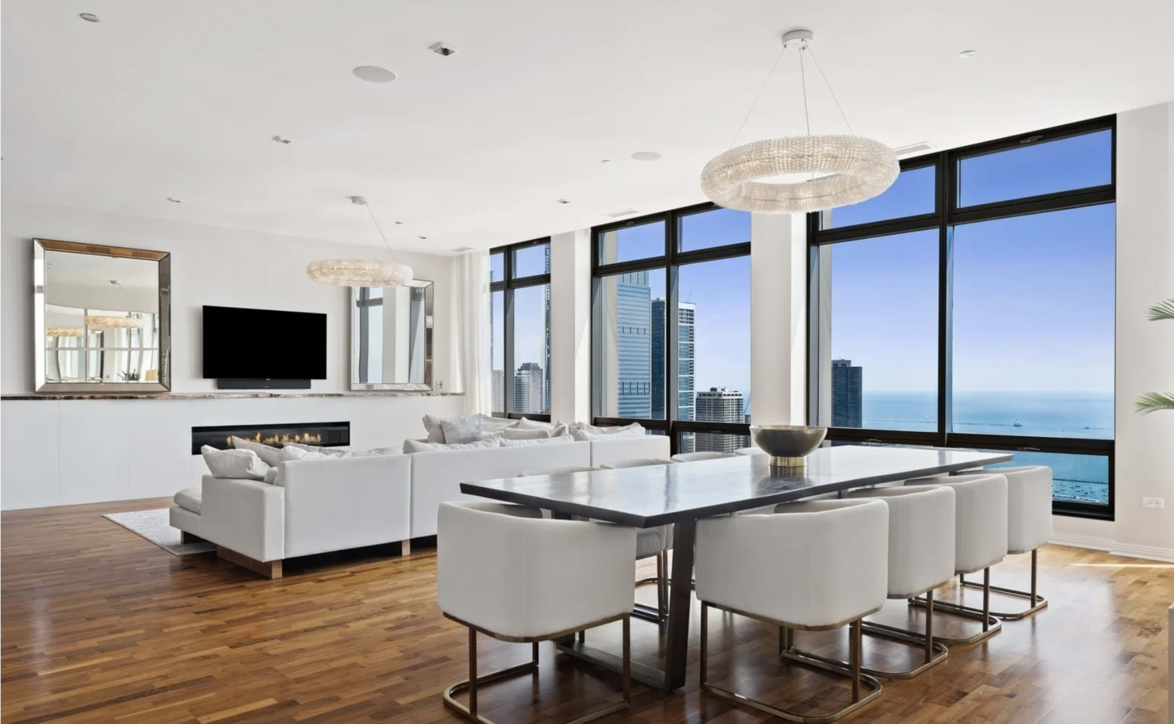 The image shows a modern living space featuring large windows with views of the ocean, a sleek dining table surrounded by white chairs, and a cozy seating area with a fireplace.