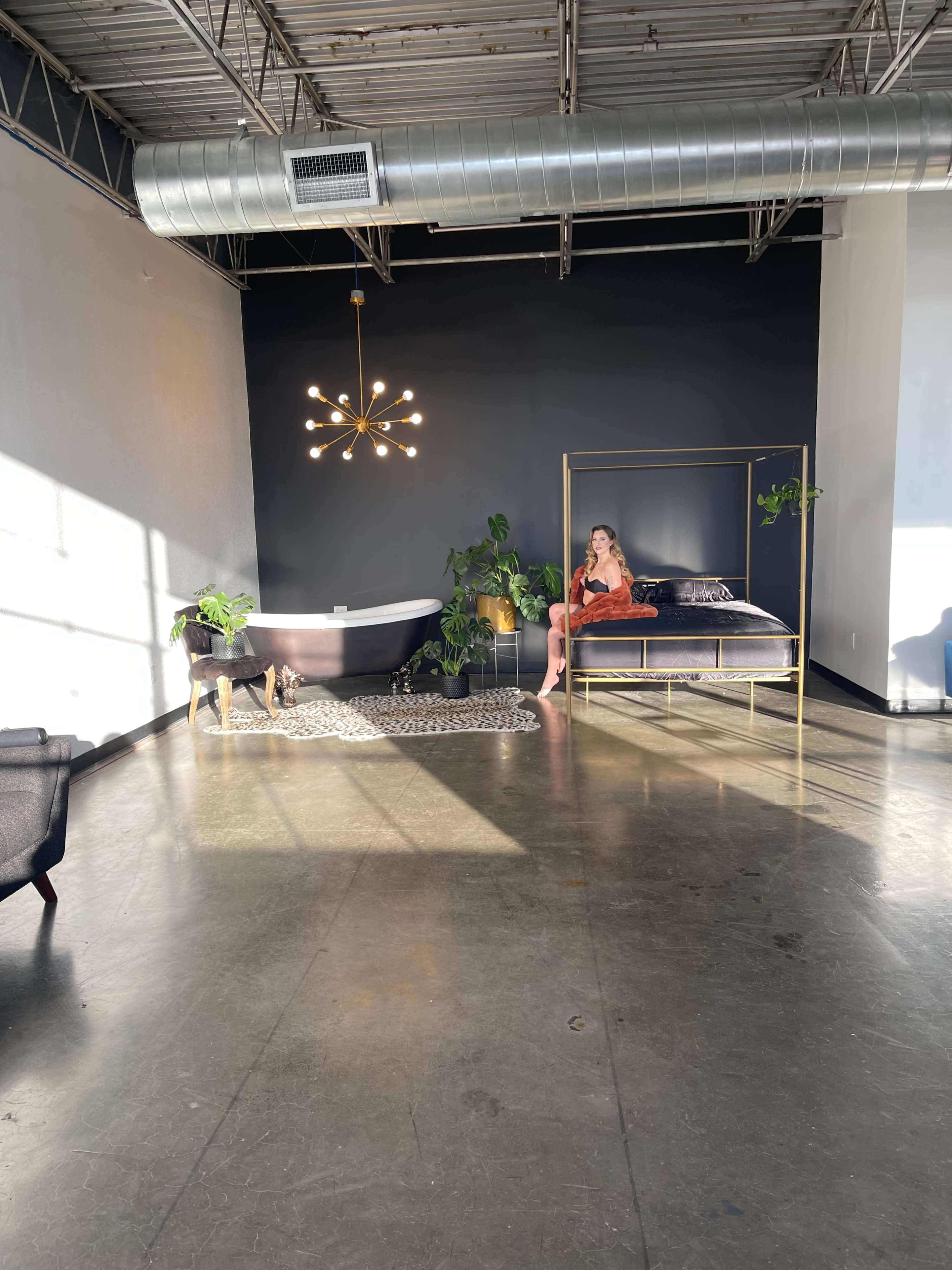 A woman sits on a bed in a spacious room featuring a freestanding bathtub, plants, and a modern light fixture.