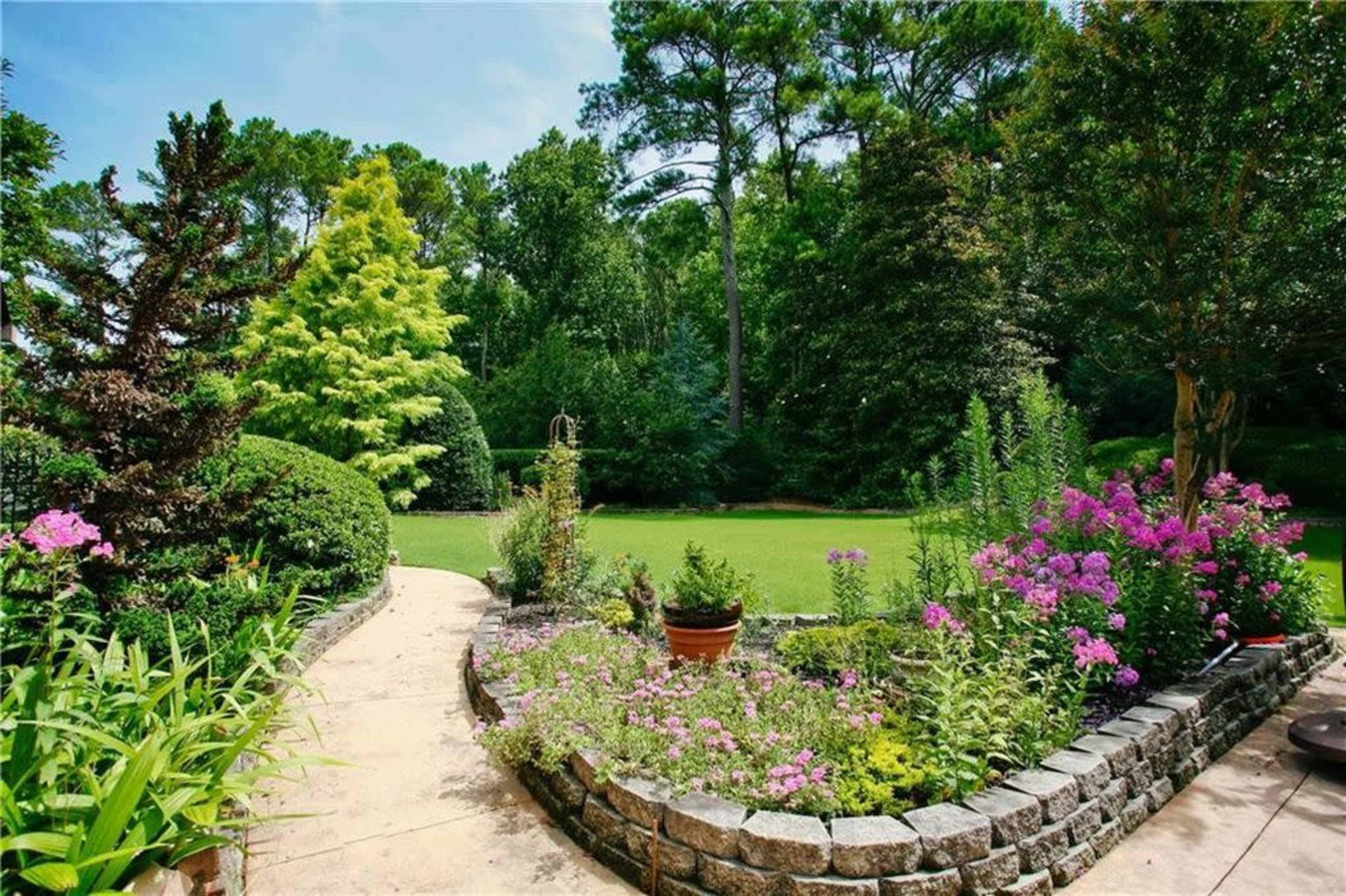 A landscaped garden features a stone border with colorful flowers and lush greenery, surrounded by trees and a clear sky.