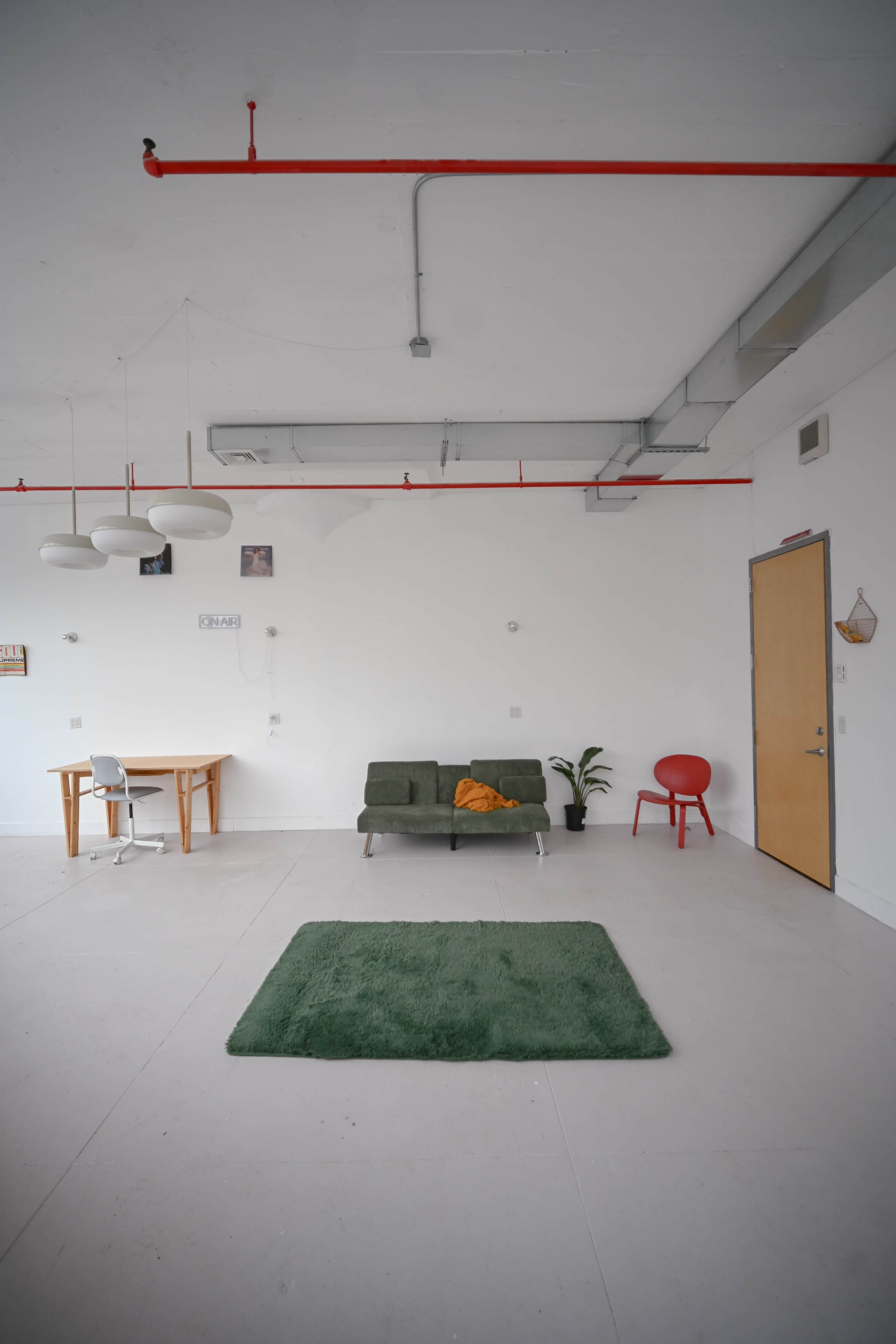 The image shows a spacious room with a green rug in the center, a green couch with an orange throw pillow, a wooden table with a chair, and a red chair against a white wall.