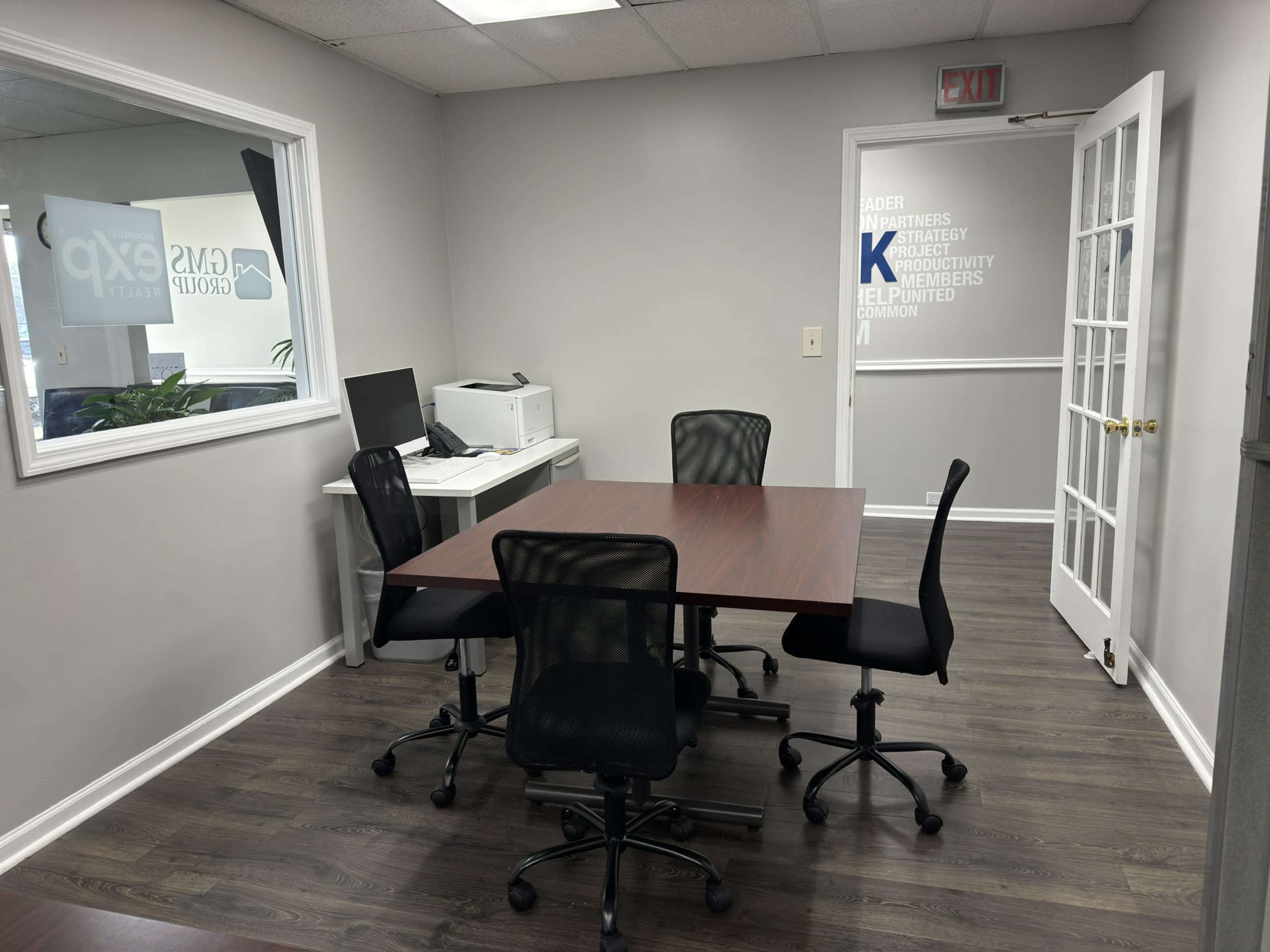 A modern office space featuring a rectangular wooden table surrounded by four rolling chairs, a computer workstation, and a printer against a wall.