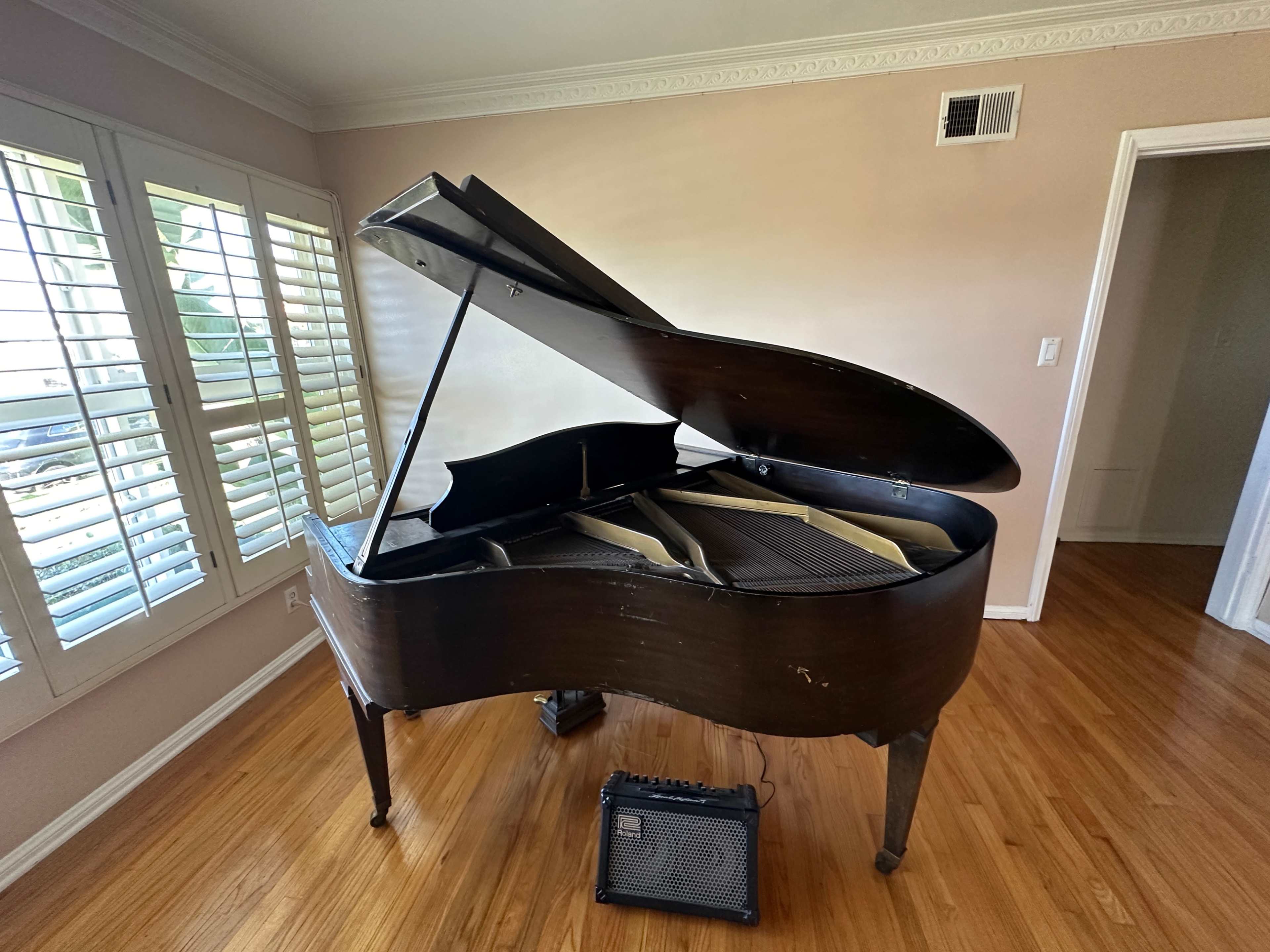 A grand piano with the lid open is positioned in a room with hardwood floors and windows featuring shuttered blinds.