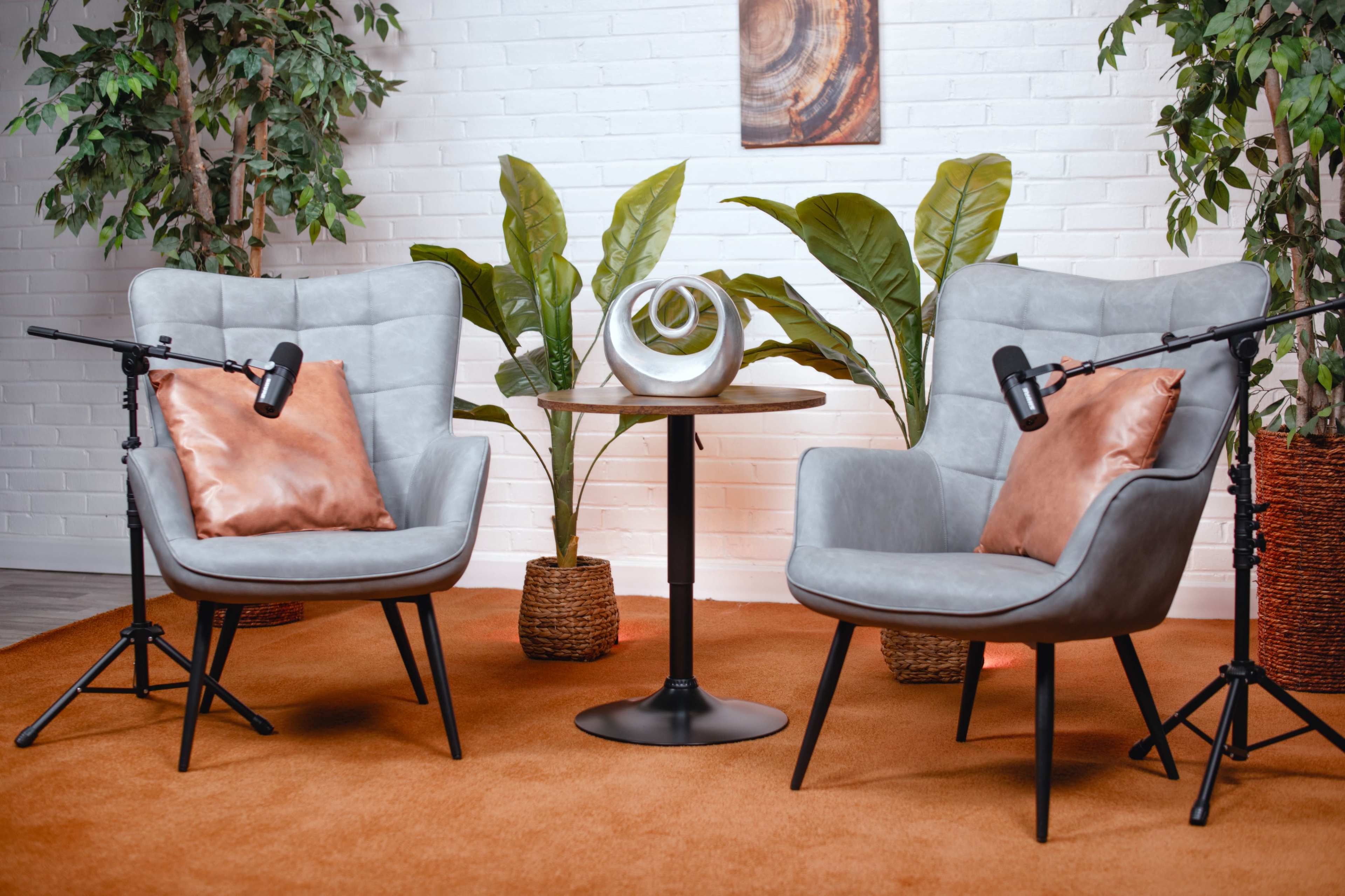 The image features two gray armchairs with orange pillows positioned near a round table, surrounded by potted plants and a vibrant orange carpet.