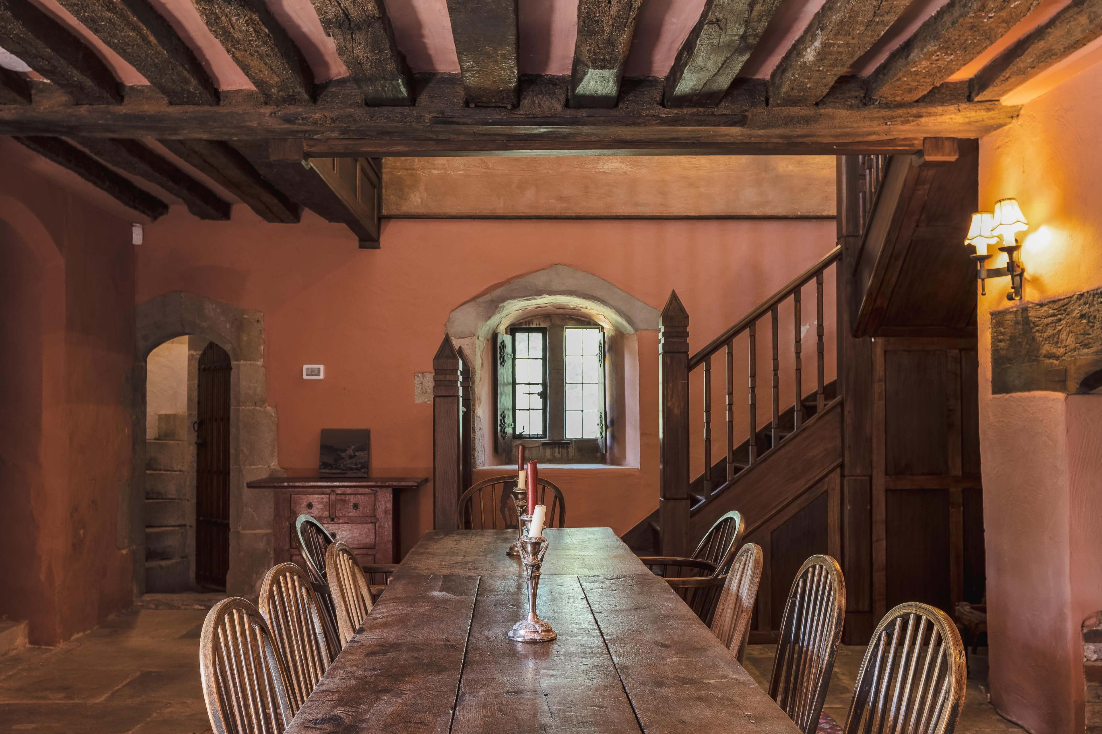 A rustic dining room featuring a long wooden table surrounded by several chairs, with exposed beams, a staircase, and softly lit walls.