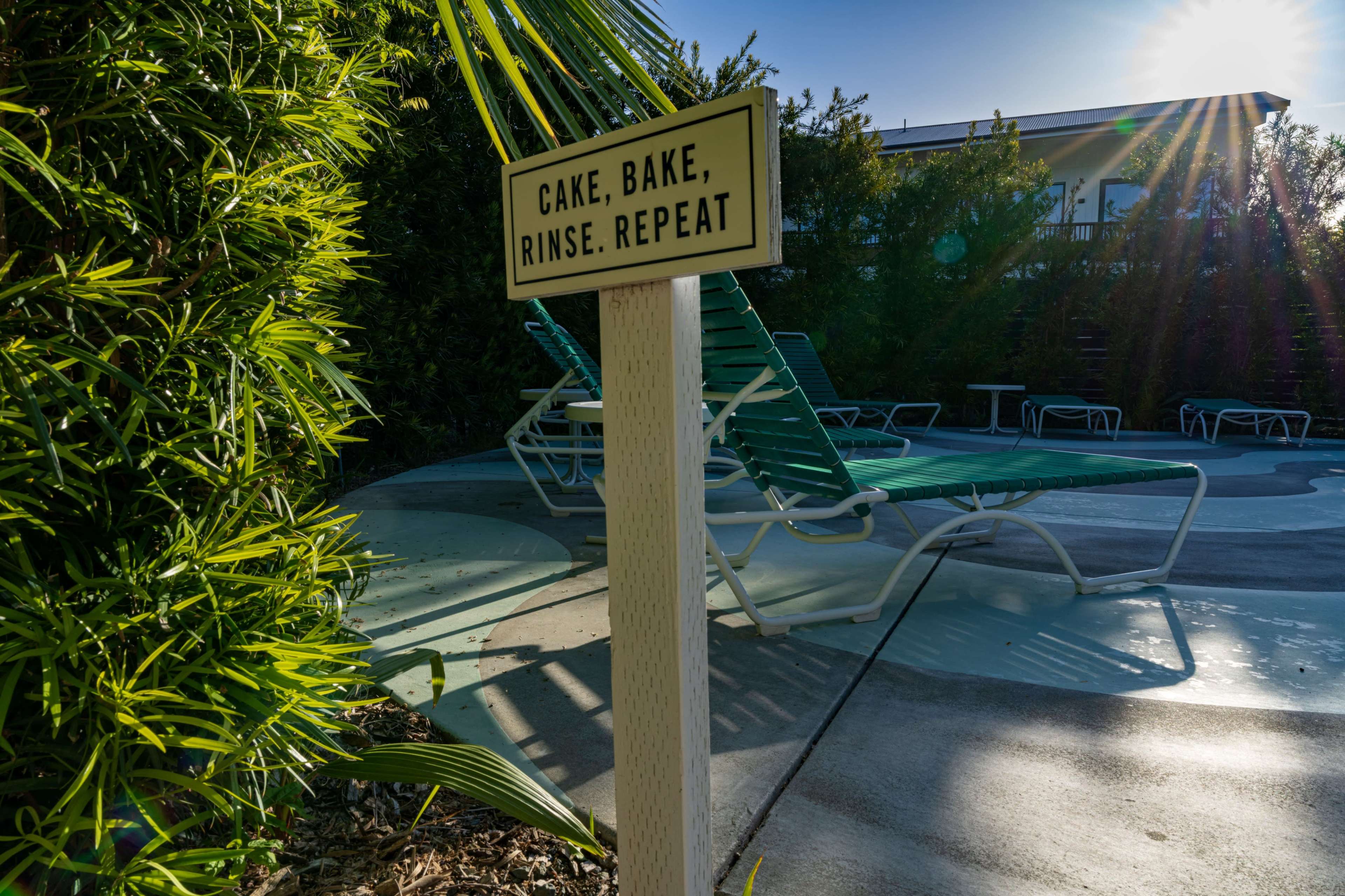 A sign reading "CAKE, BAKE, RINSE, REPEAT" is positioned next to a pool area with lounge chairs and lush greenery.