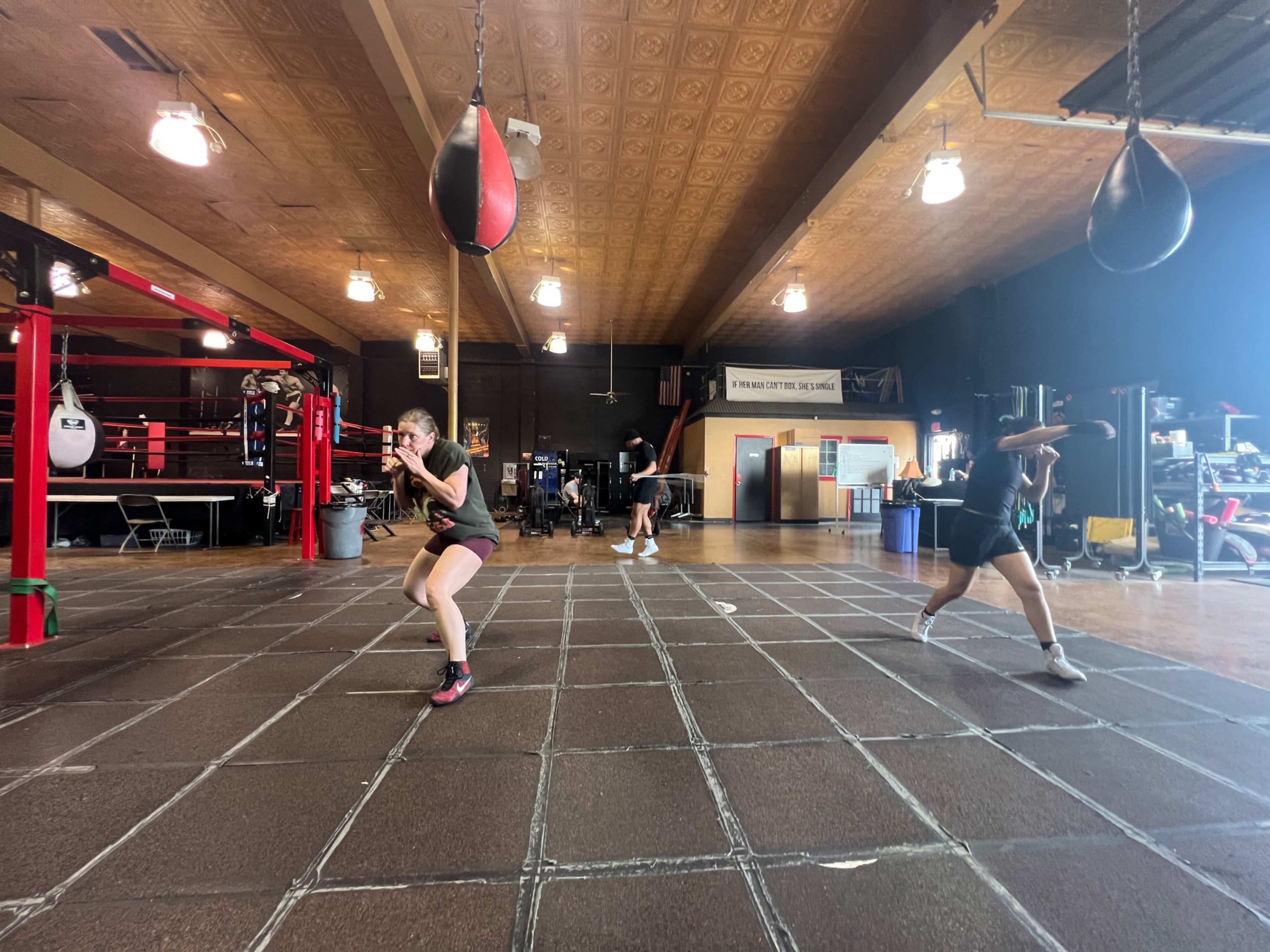 Two individuals are training in a boxing gym, with one person practicing on a heavy bag and another shadow boxing in the foreground.