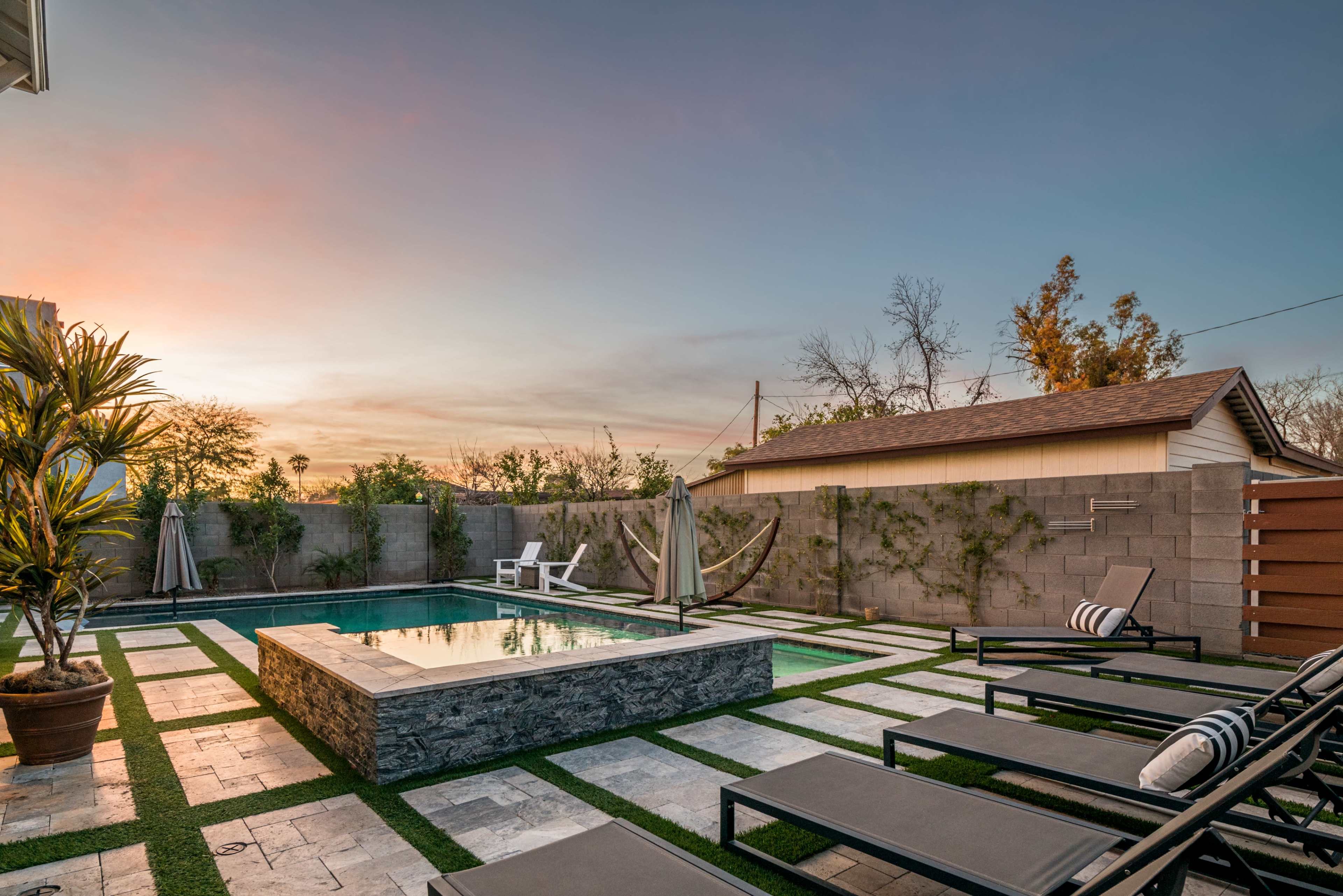 A backyard scene features a swimming pool surrounded by lounge chairs and landscaped with stone tiles and greenery under a sunset sky.