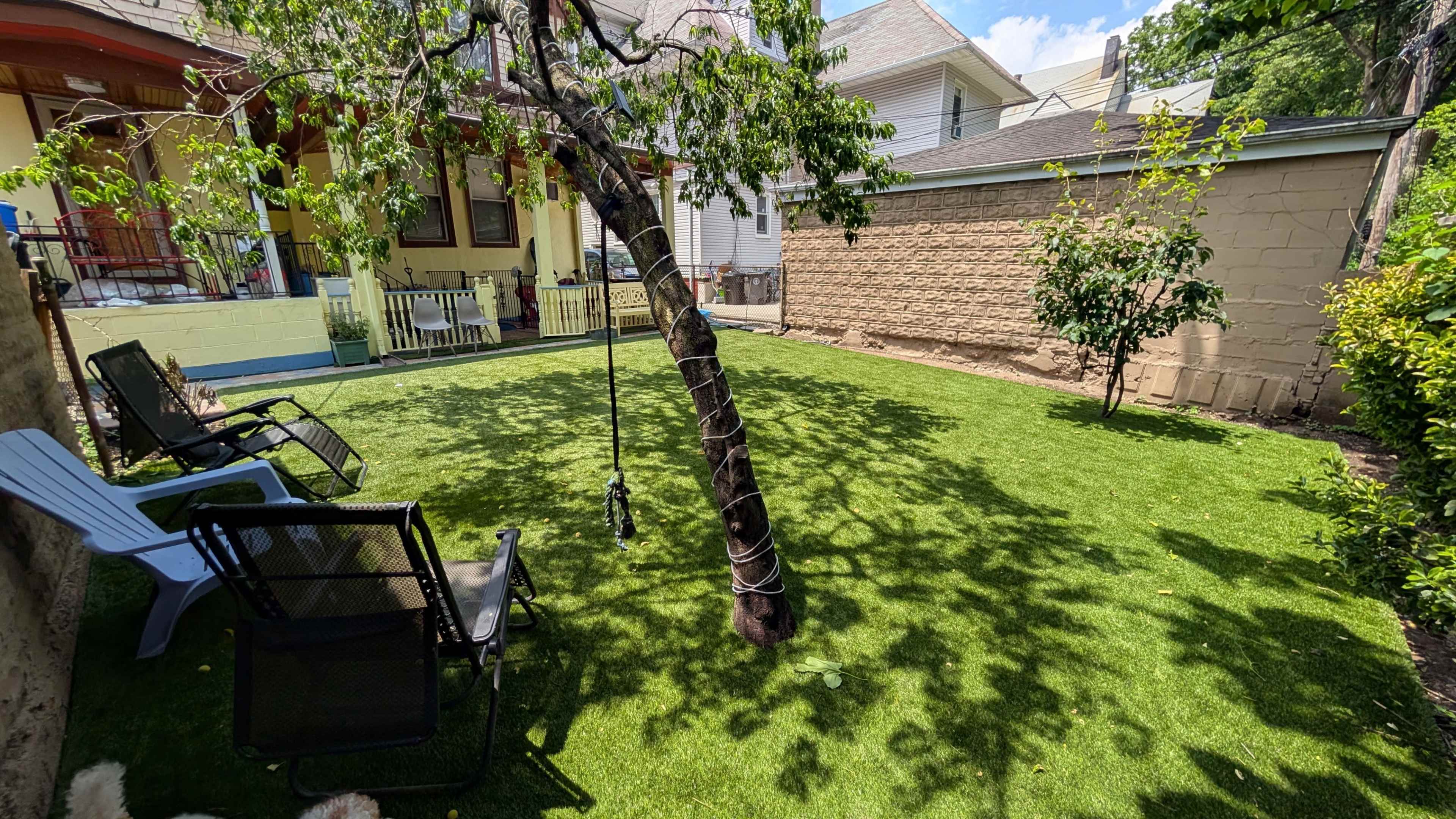 A green lawn with artificial turf features a tree with a swing, surrounded by chairs and buildings in a residential yard.