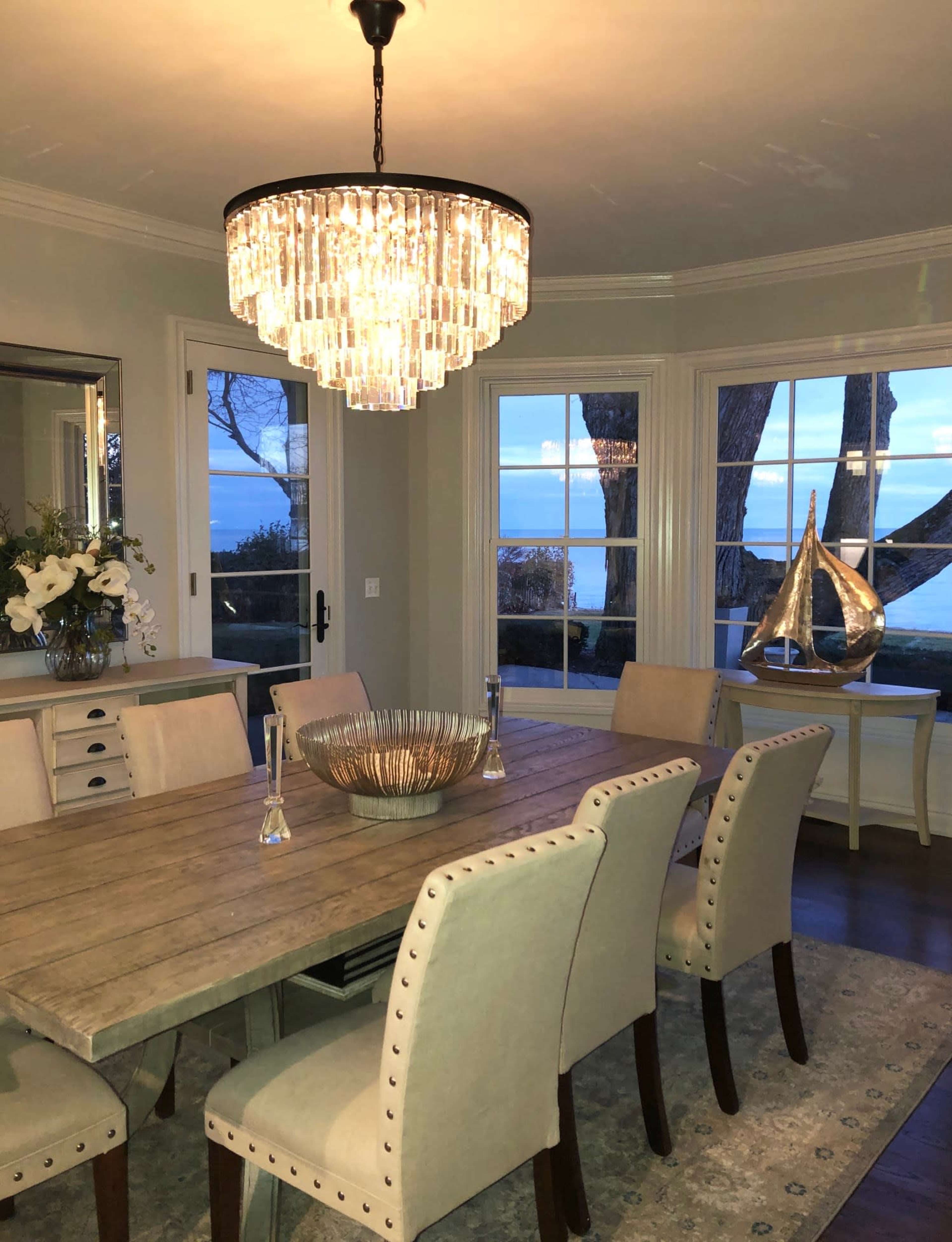 A dining room features a wooden table surrounded by upholstered chairs, a crystal chandelier overhead, and large windows displaying a view of the water.