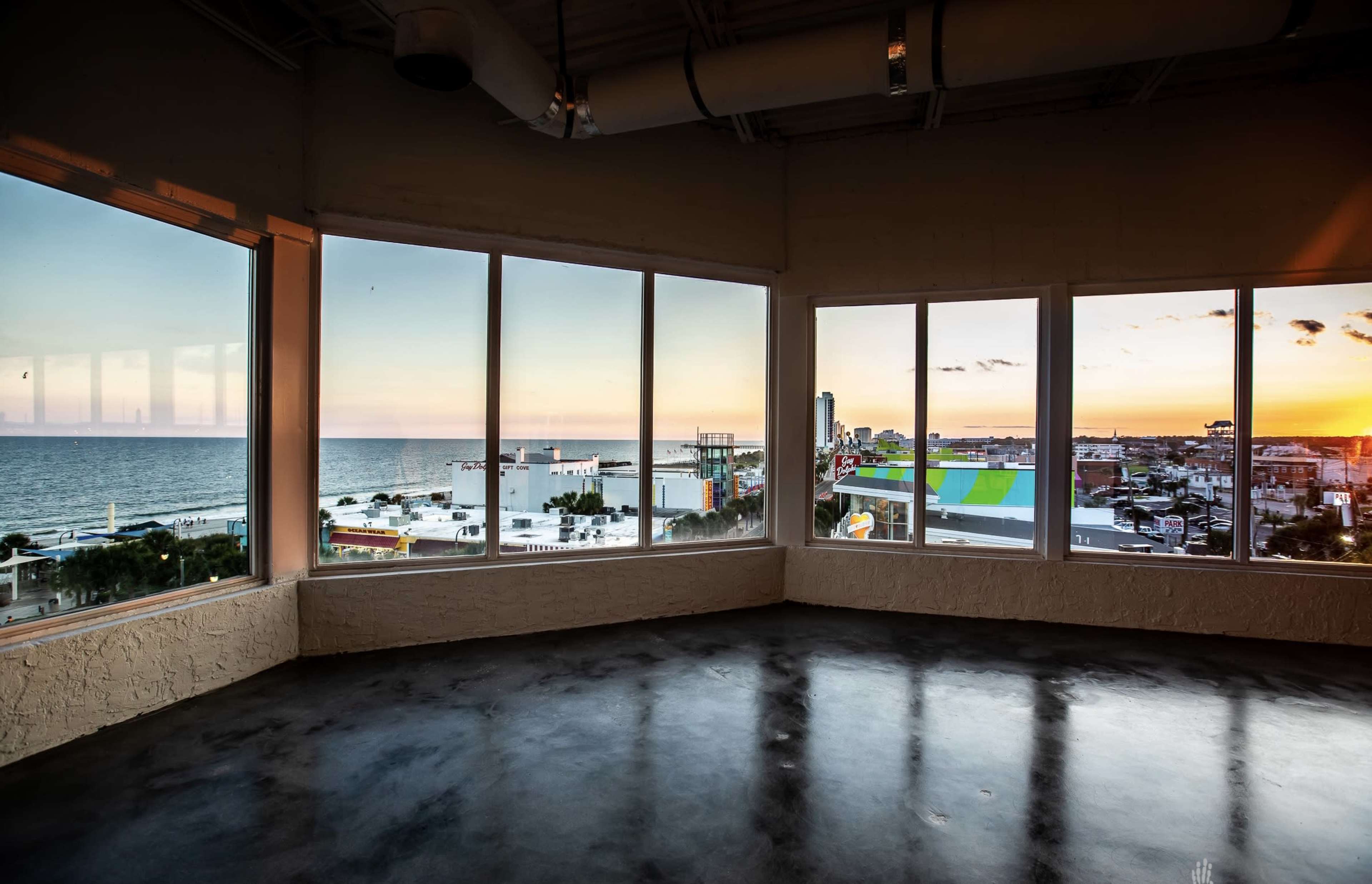 A spacious, modern room with large windows overlooking a coastal sunset and cityscape.