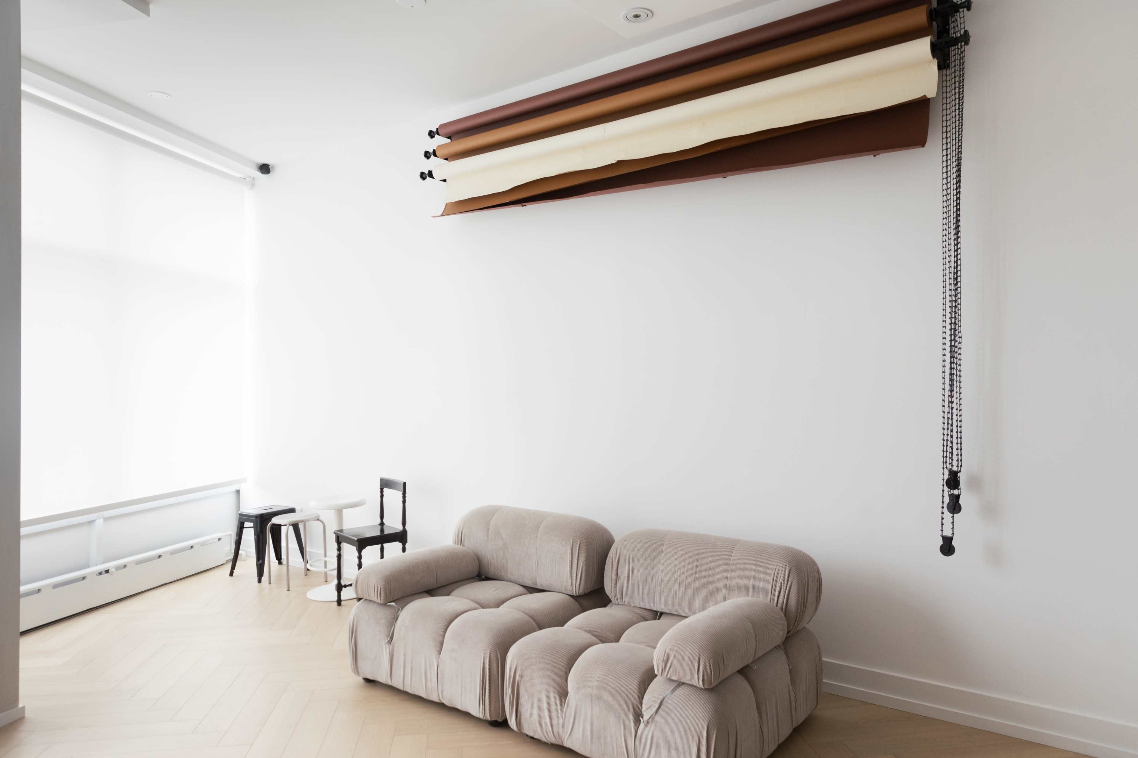 The image shows a minimalistic interior featuring a light-colored wall, a beige sofa, two small tables, and a roll of backdrop paper mounted on the wall.