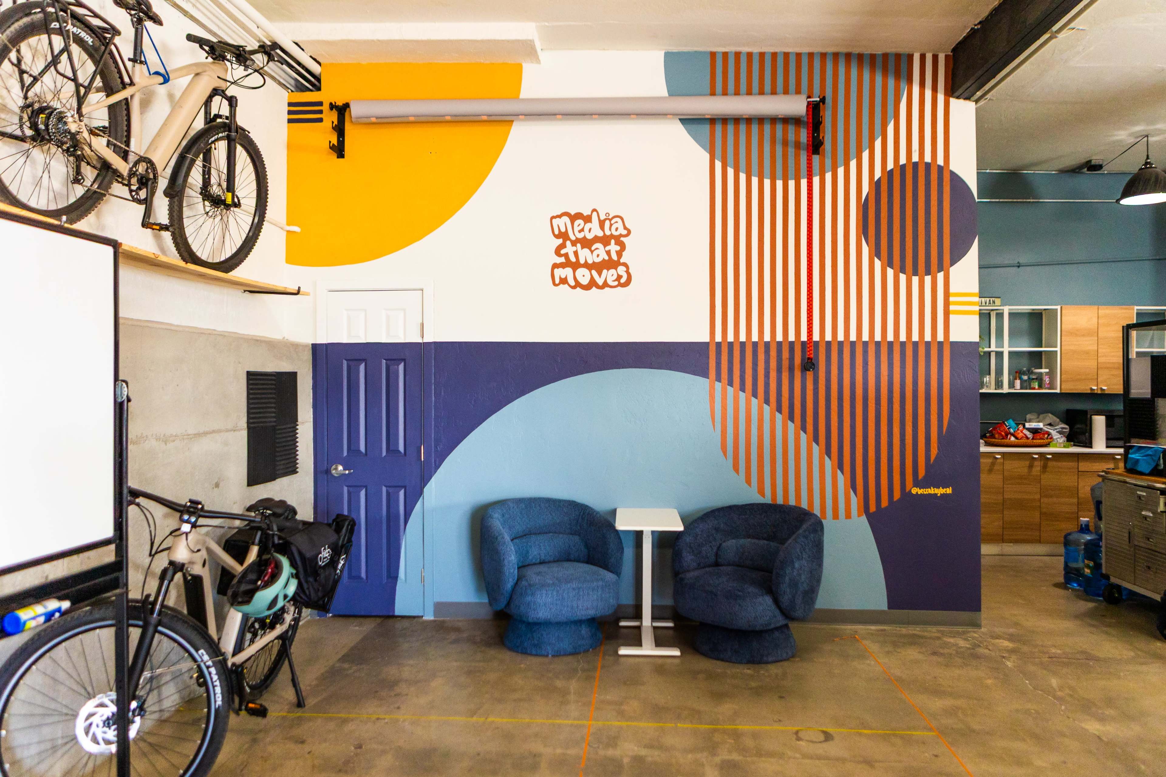 The image shows a garage-style workspace with two blue chairs, a painted wall featuring colorful geometric designs, and bicycles mounted on a shelf above.
