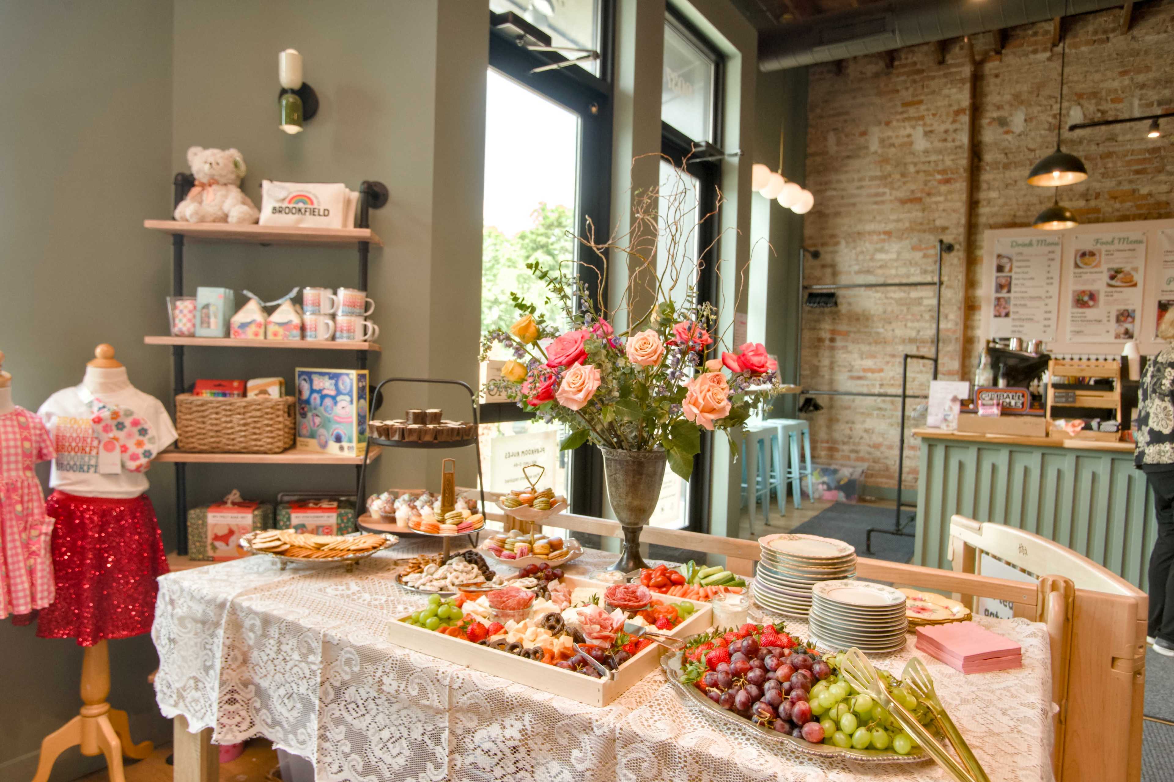 A decorated table showcases a variety of food items, including fruits, pastries, and a floral arrangement, in a cafe setting.