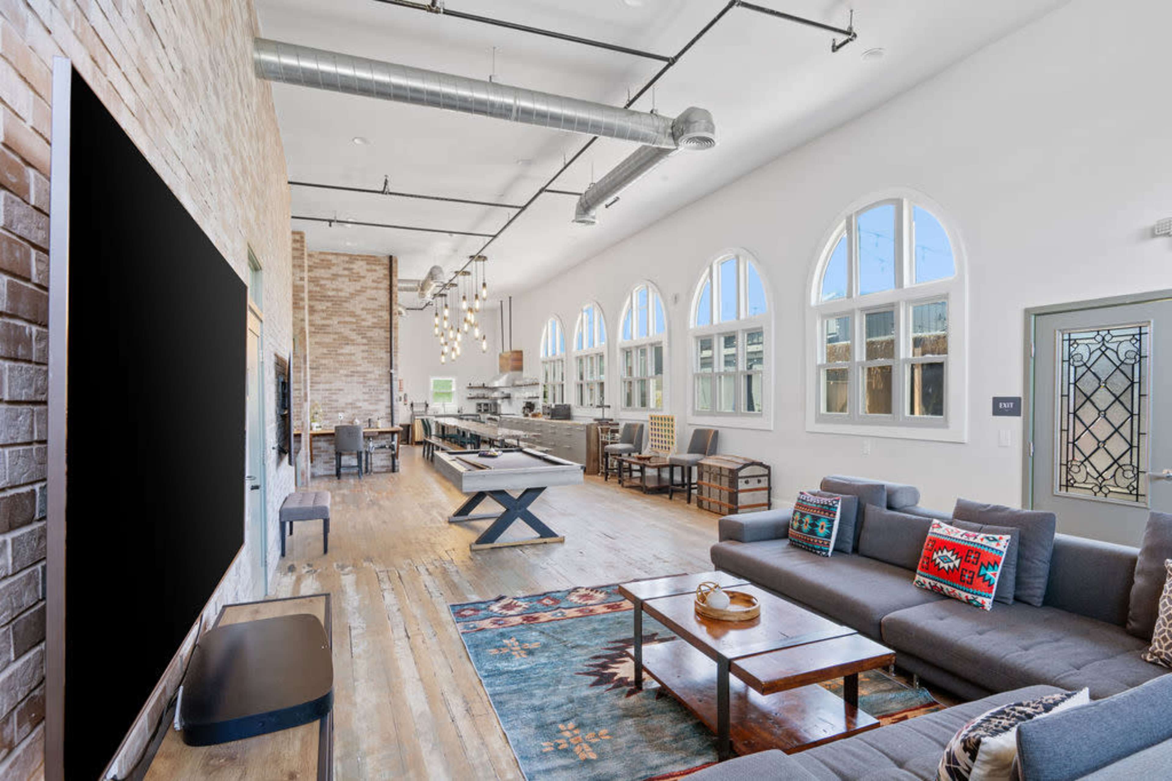 The image shows an open-concept living space with high ceilings, large arched windows, a modern kitchen, and a comfortable seating area featuring a dark sofa and a wooden coffee table.