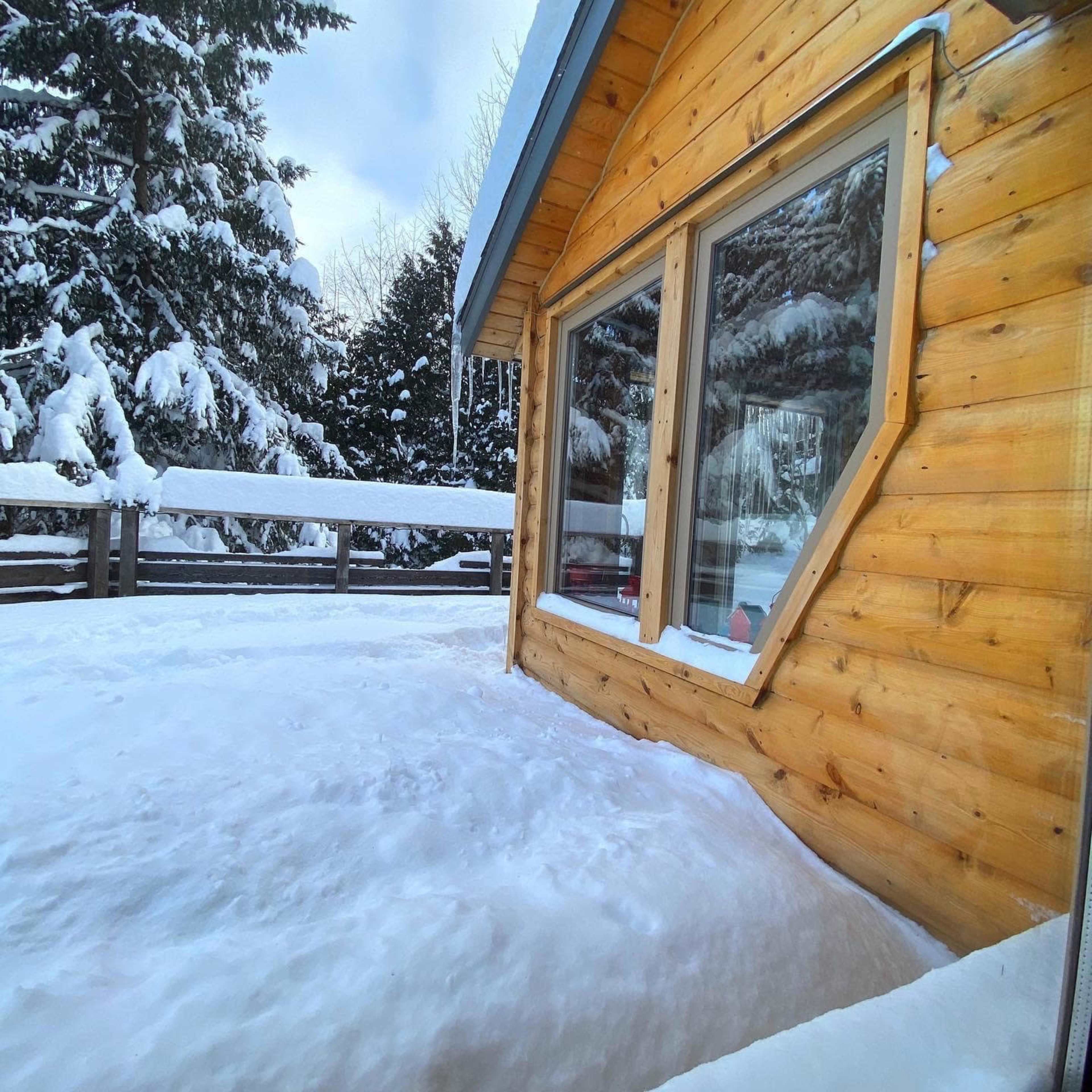 A wooden cabin sits amidst deep snow, with snow-covered trees in the background and icicles hanging from the roof.