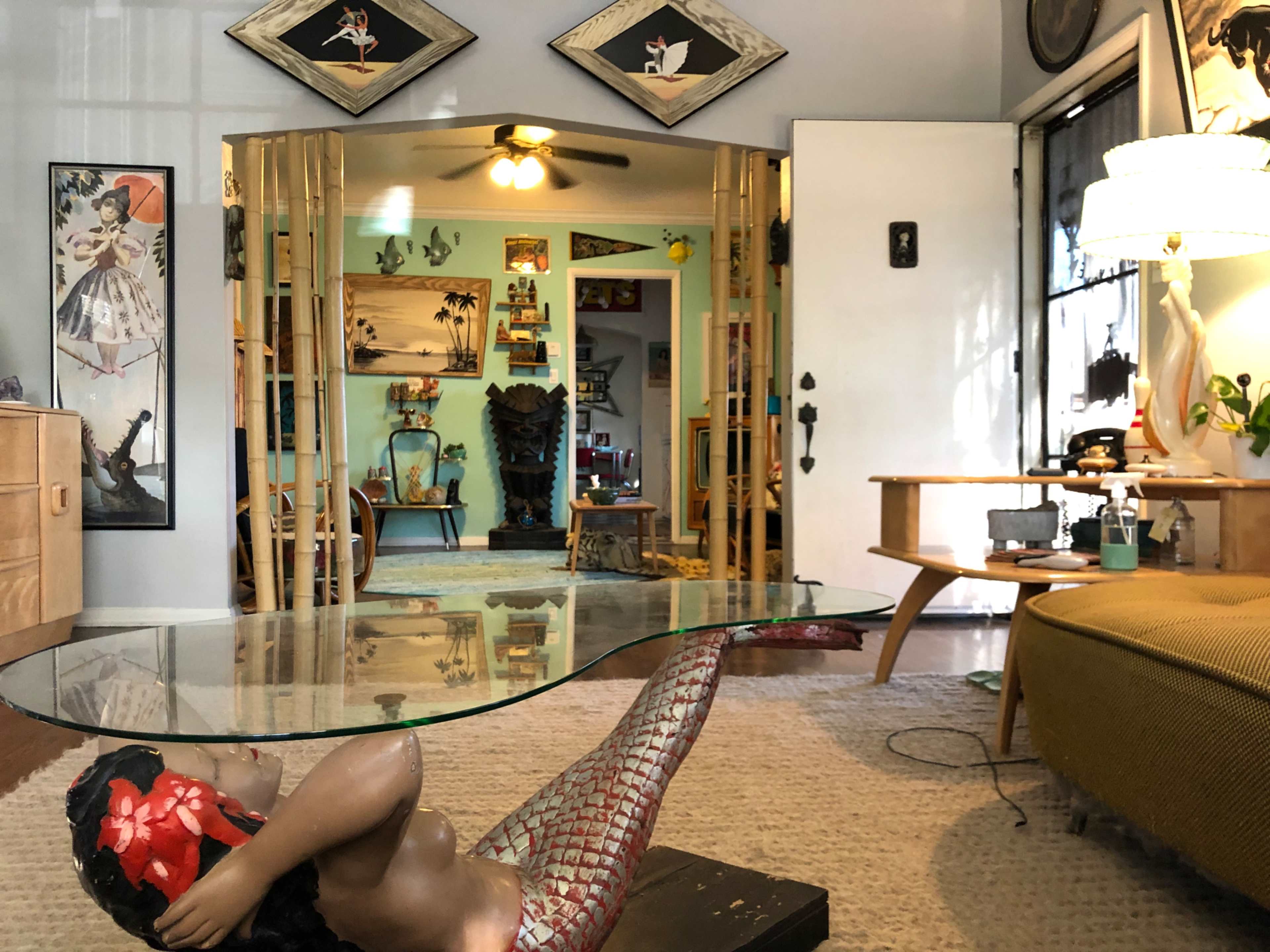 The image features a vintage living room with a glass coffee table supported by a mermaid figure, framed artwork on the walls, and a bright green wall in the background.
