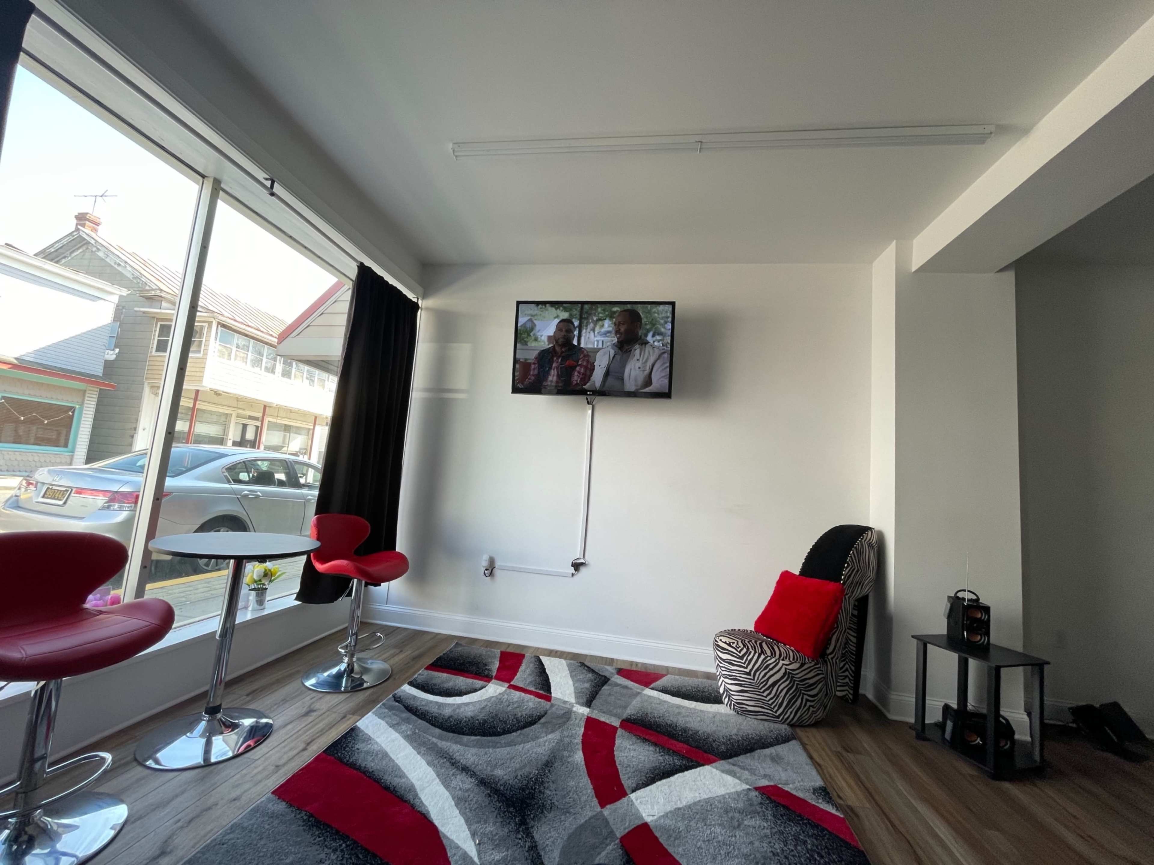 A modern, well-lit room featuring two red barstools, a zebra-striped chair, a large TV mounted on the wall, and a patterned gray rug on the floor.