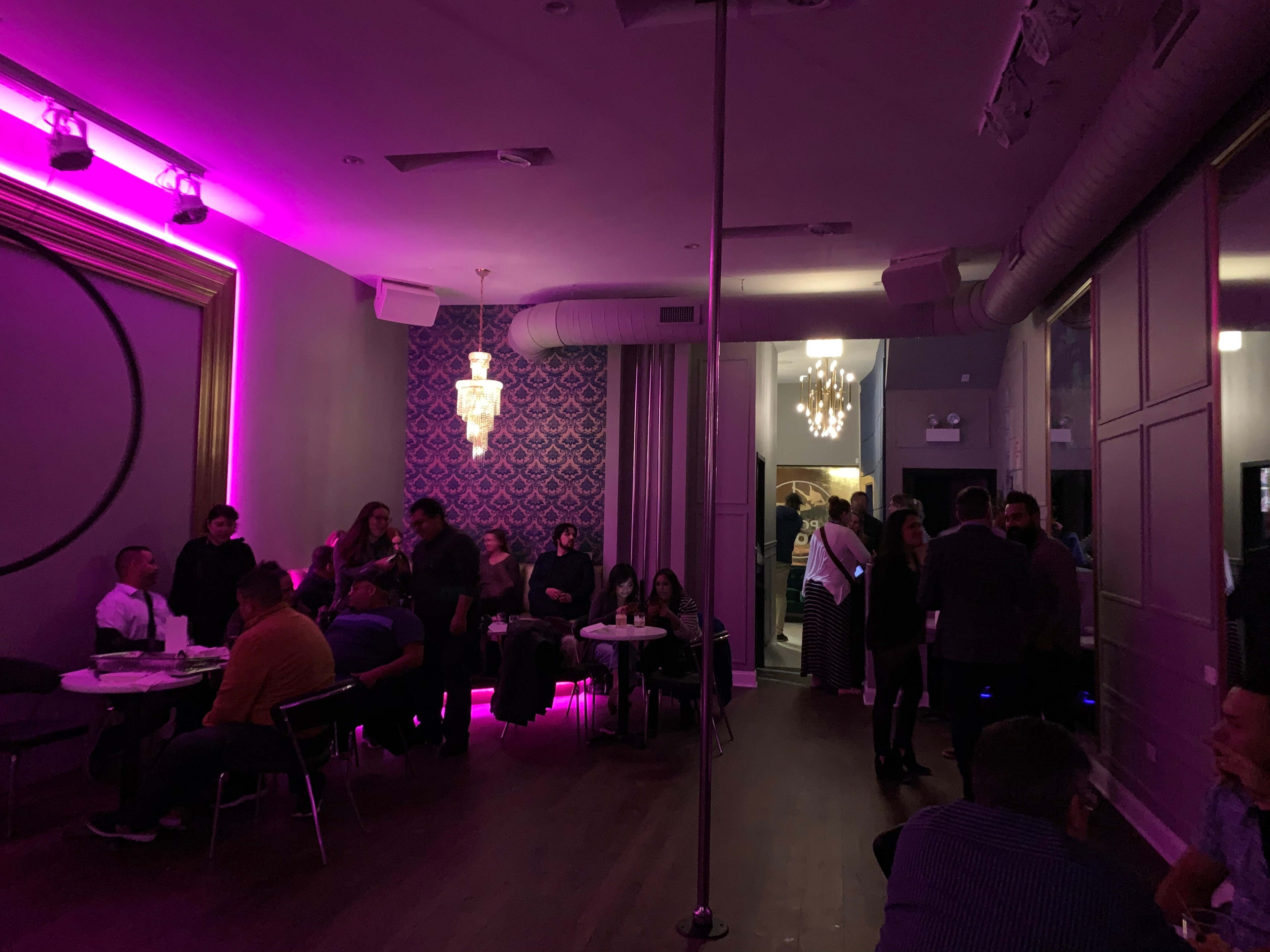 A lively indoor venue with pink lighting, featuring clusters of people socializing at tables and a pole in the center of the room.