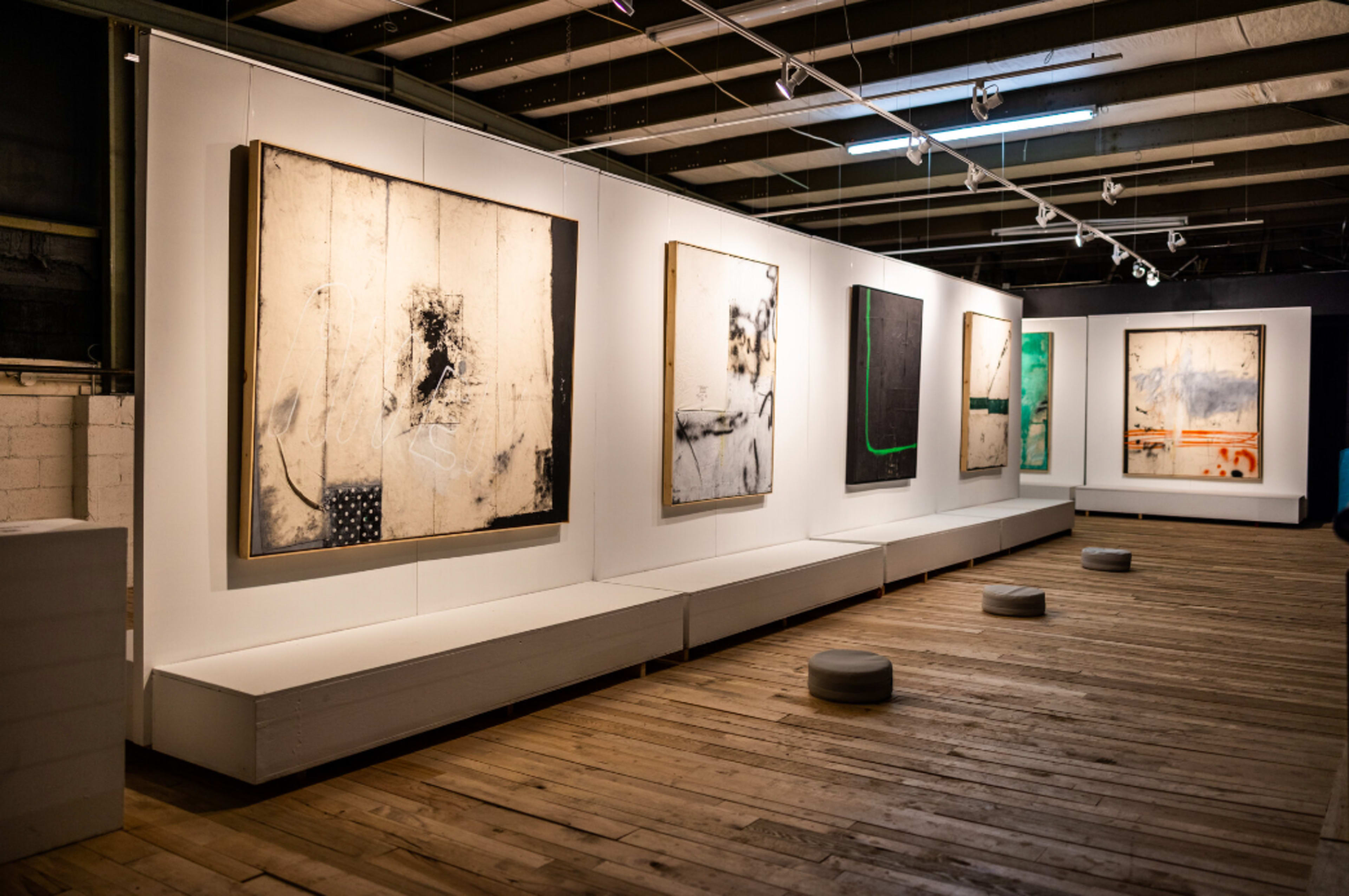 The image shows an art gallery featuring a series of abstract paintings displayed on white walls with wooden flooring.