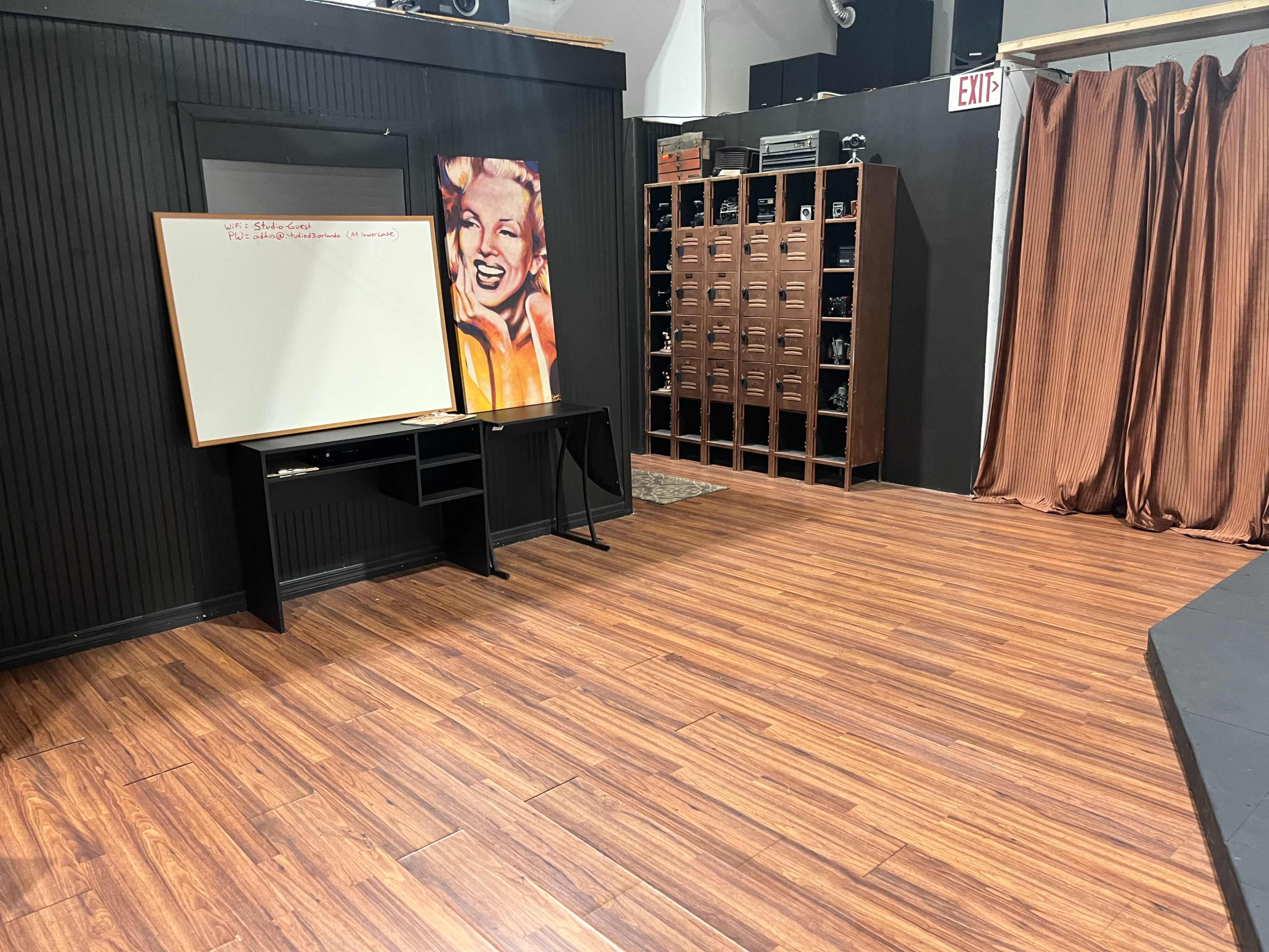 The image shows a rehearsal space with a whiteboard, a black table, and wooden lockers against a wall, featuring a portrait of a smiling woman.