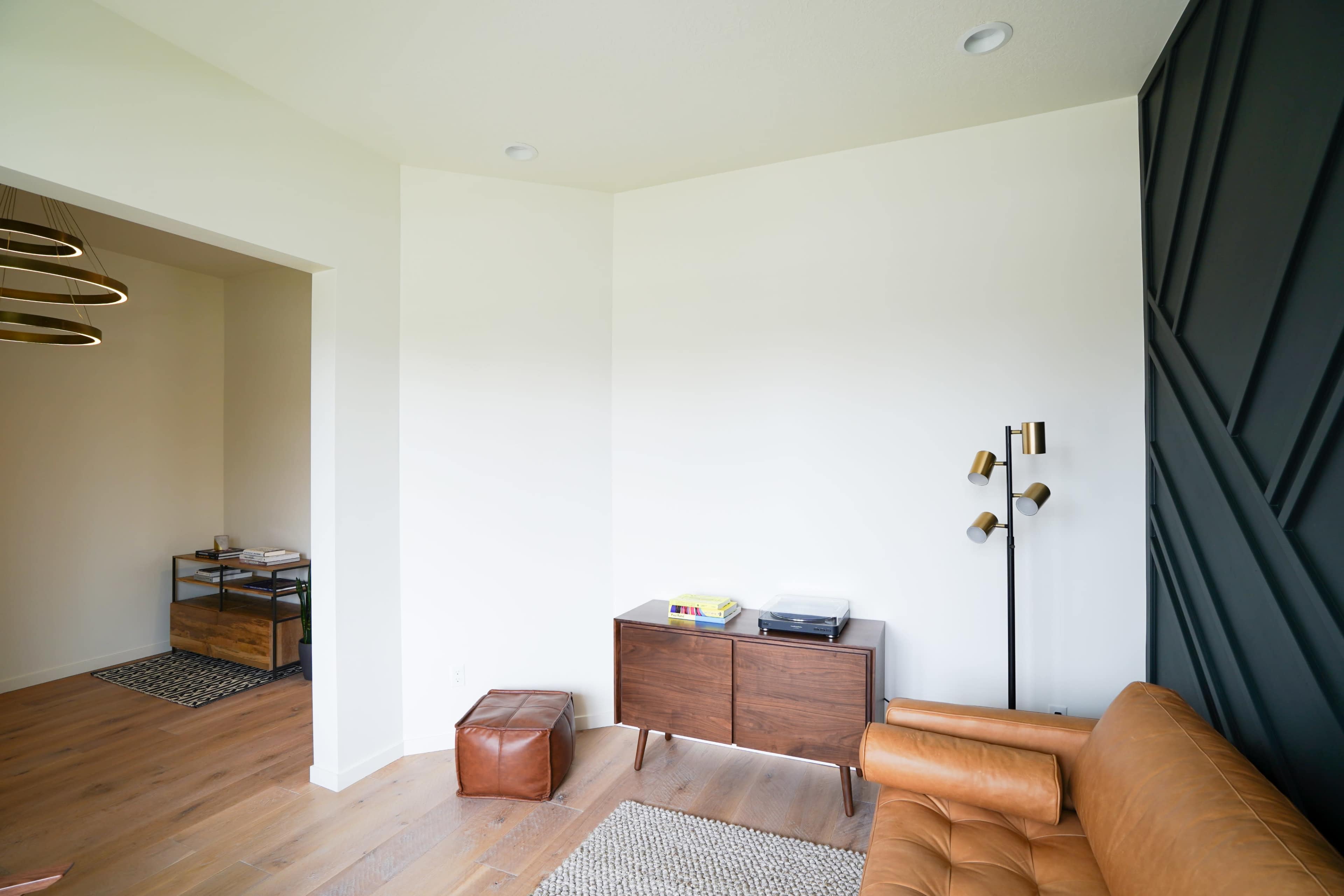 The image shows a light-filled room featuring a leather sofa, a wooden sideboard, a standing lamp, and a textured wall.