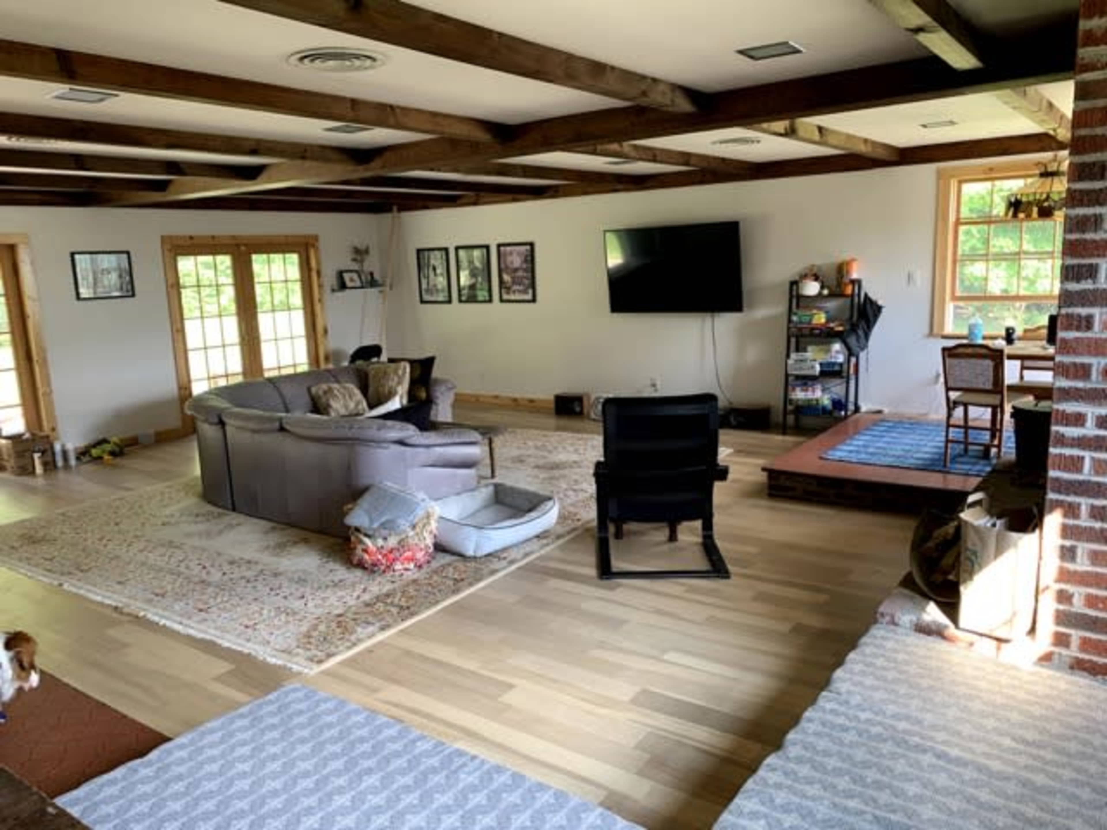 A spacious living room with wooden beams, a large sectional sofa, a television mounted on the wall, and a laid-back seating area near a window.
