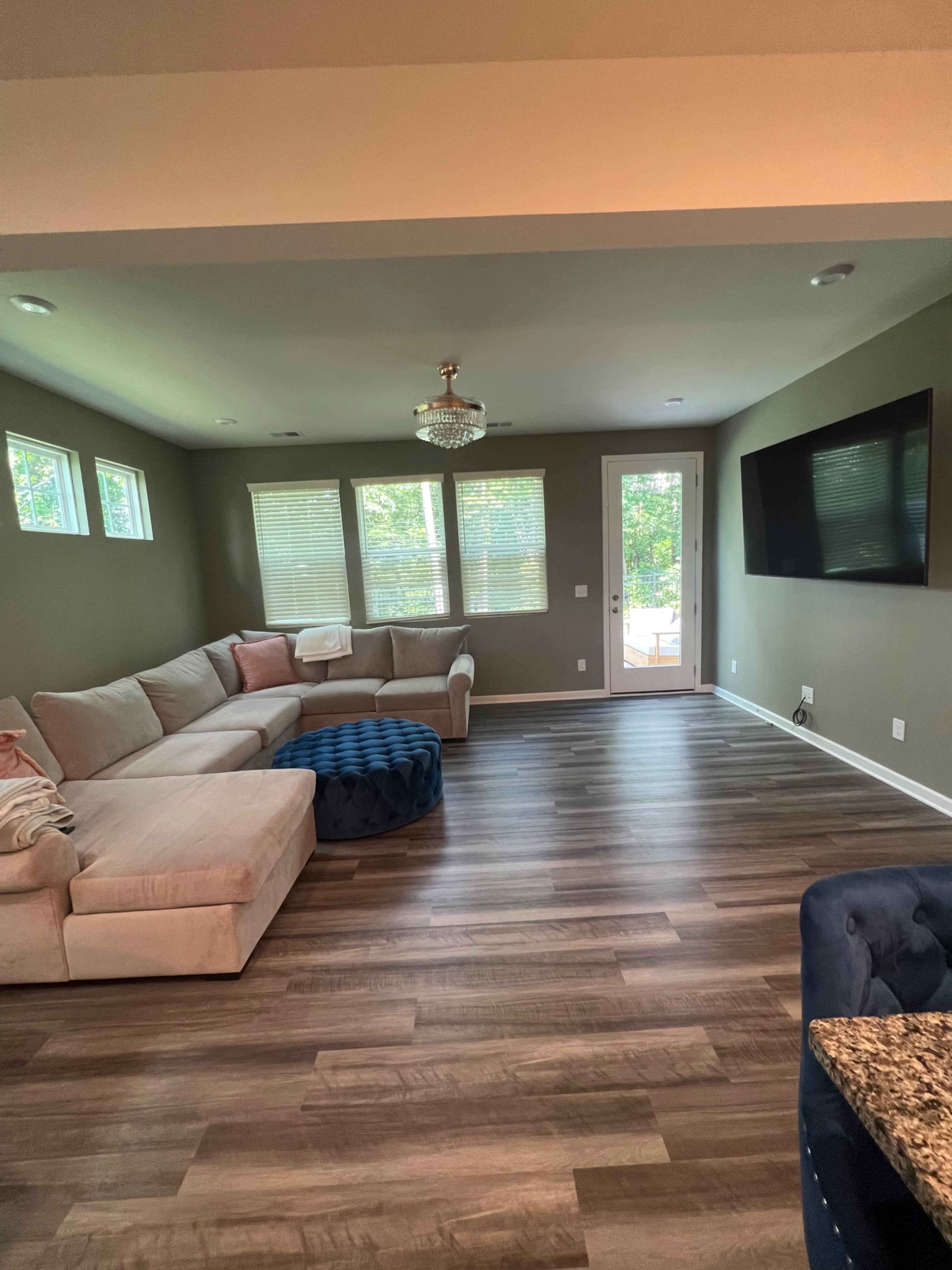 The living room features a sectional sofa, a round ottoman, a large TV, and windows that provide natural light.