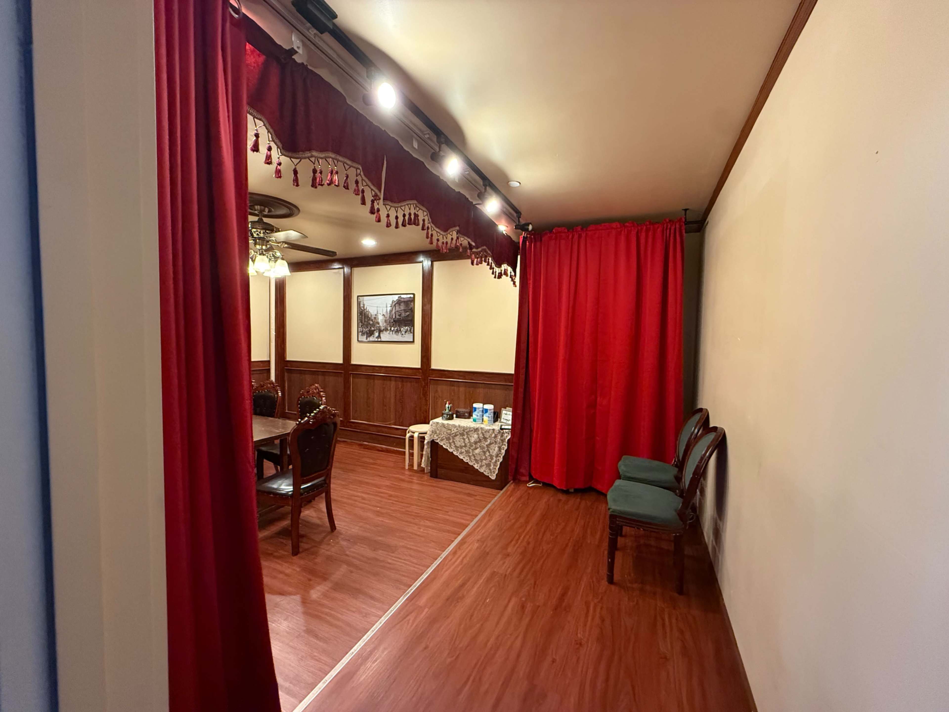 The image shows a room with a wooden floor, a dining table set with chairs, and red curtains hanging from the ceiling.