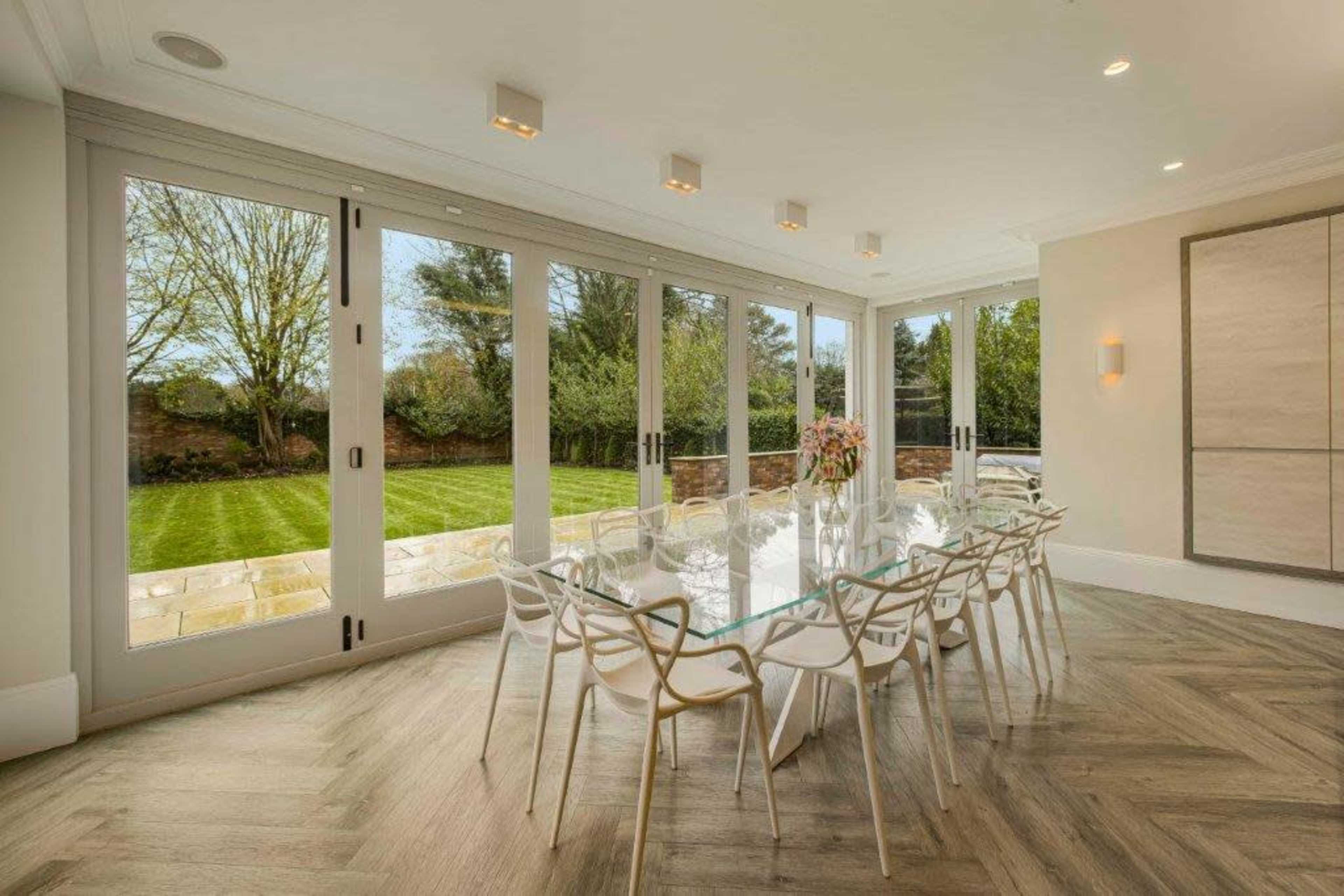 A modern dining room features a glass table and white chairs, with large windows offering a view of a grassy backyard.