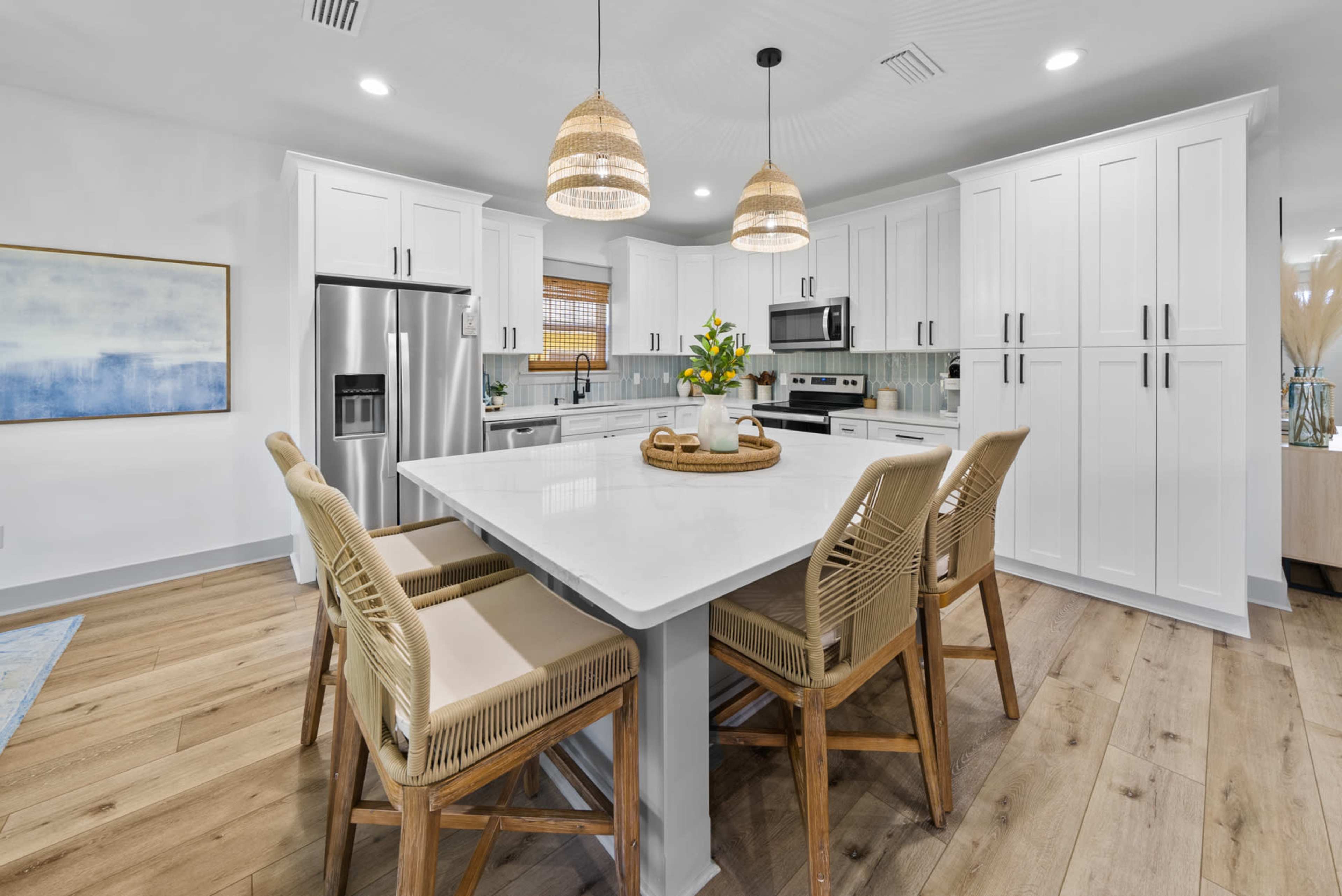 A modern kitchen features white cabinetry, stainless steel appliances, and a large island surrounded by four wicker-barred chairs.