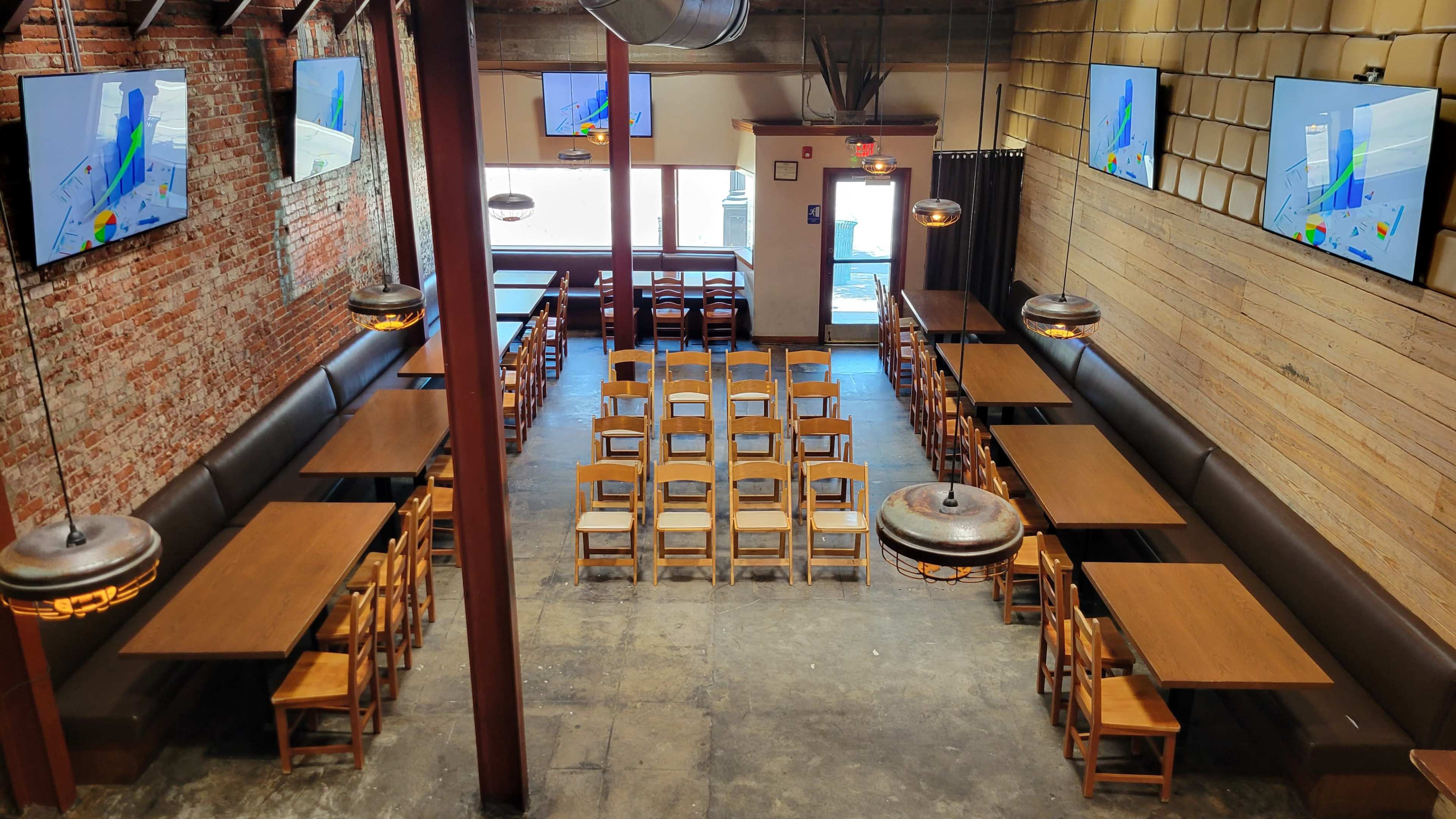 A spacious dining area with wooden tables and chairs arranged in rows, flanked by brick and wooden walls, and featuring two large screens displaying maps.