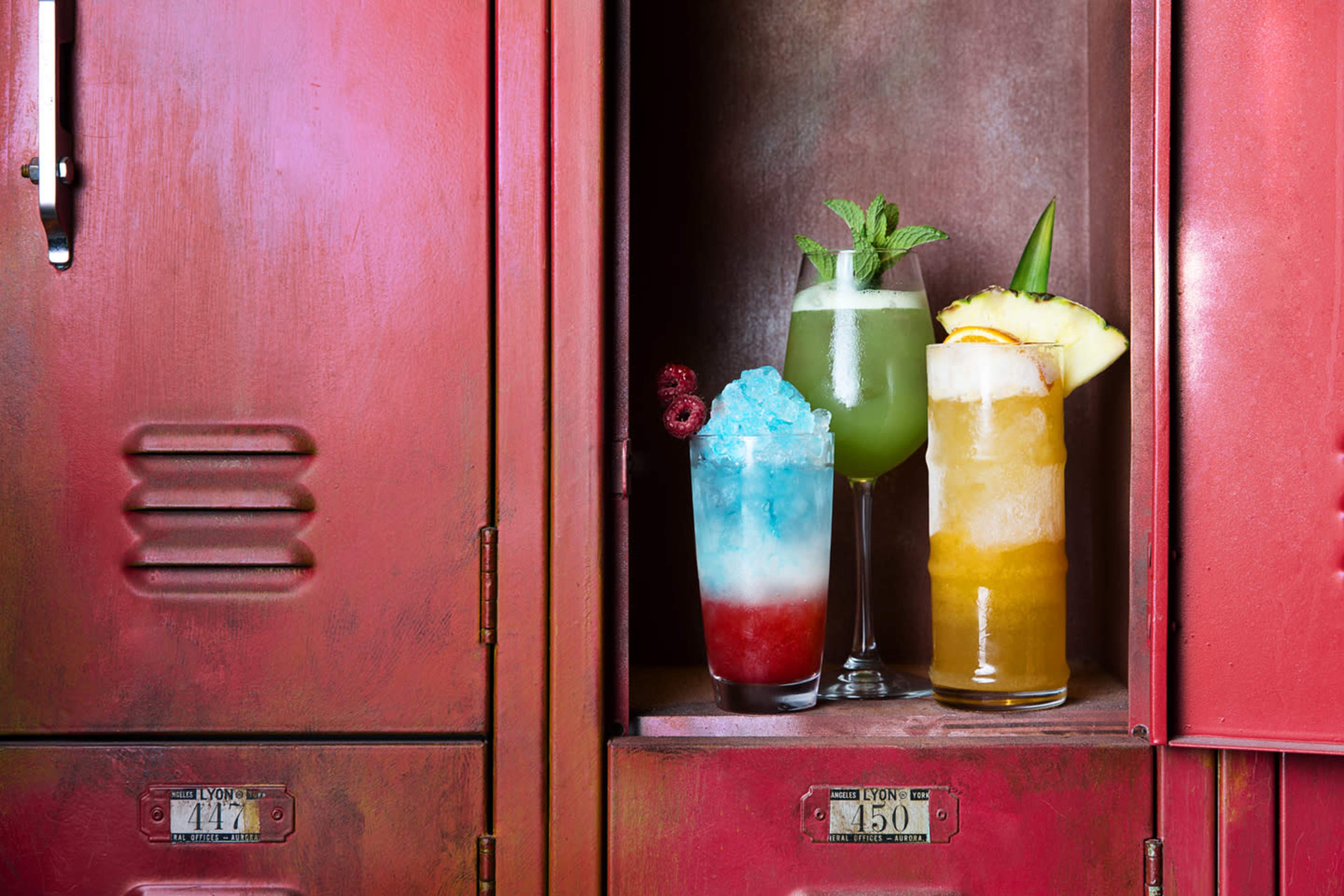 The image displays three colorful cocktails arranged on a shelf inside a red locker.