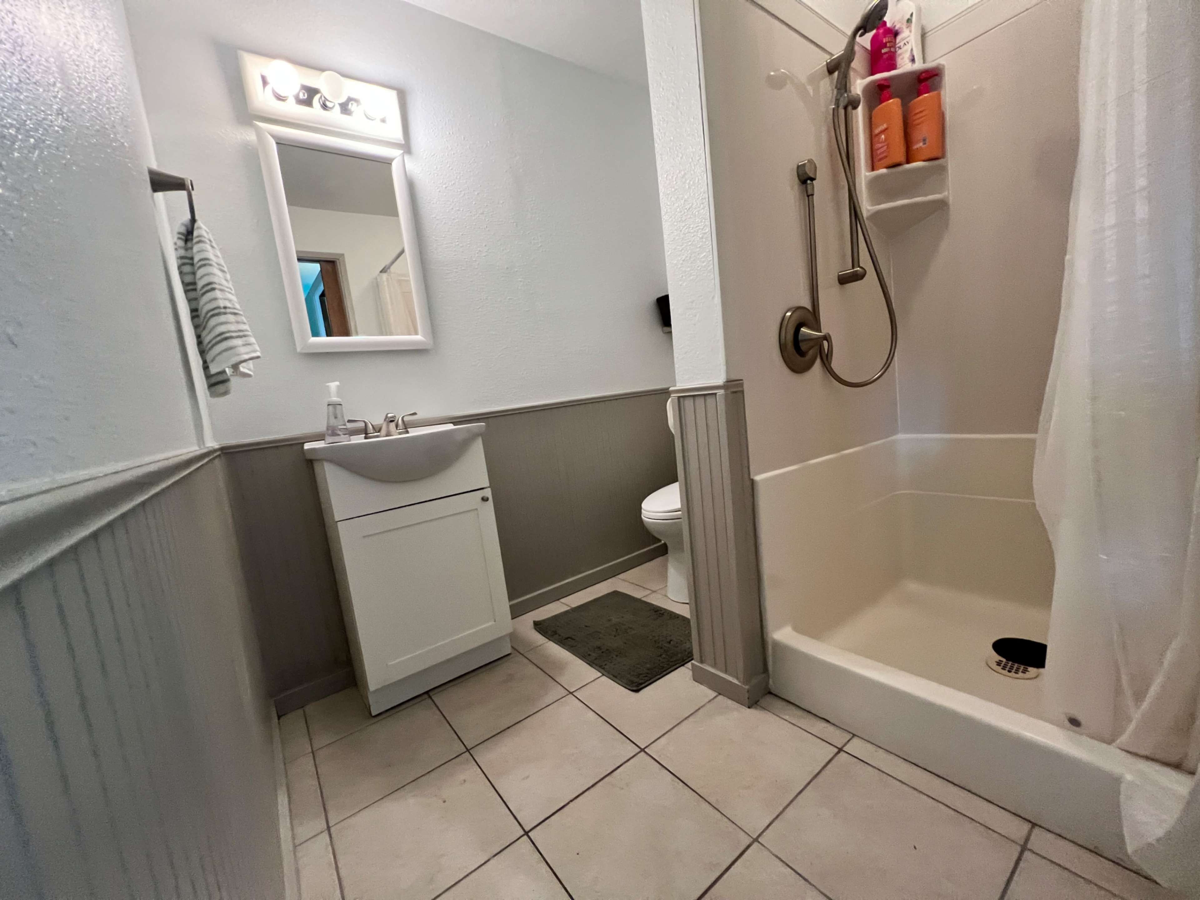 The image shows a compact bathroom featuring a shower, a sink with a mirror above it, and light-colored wall panels.