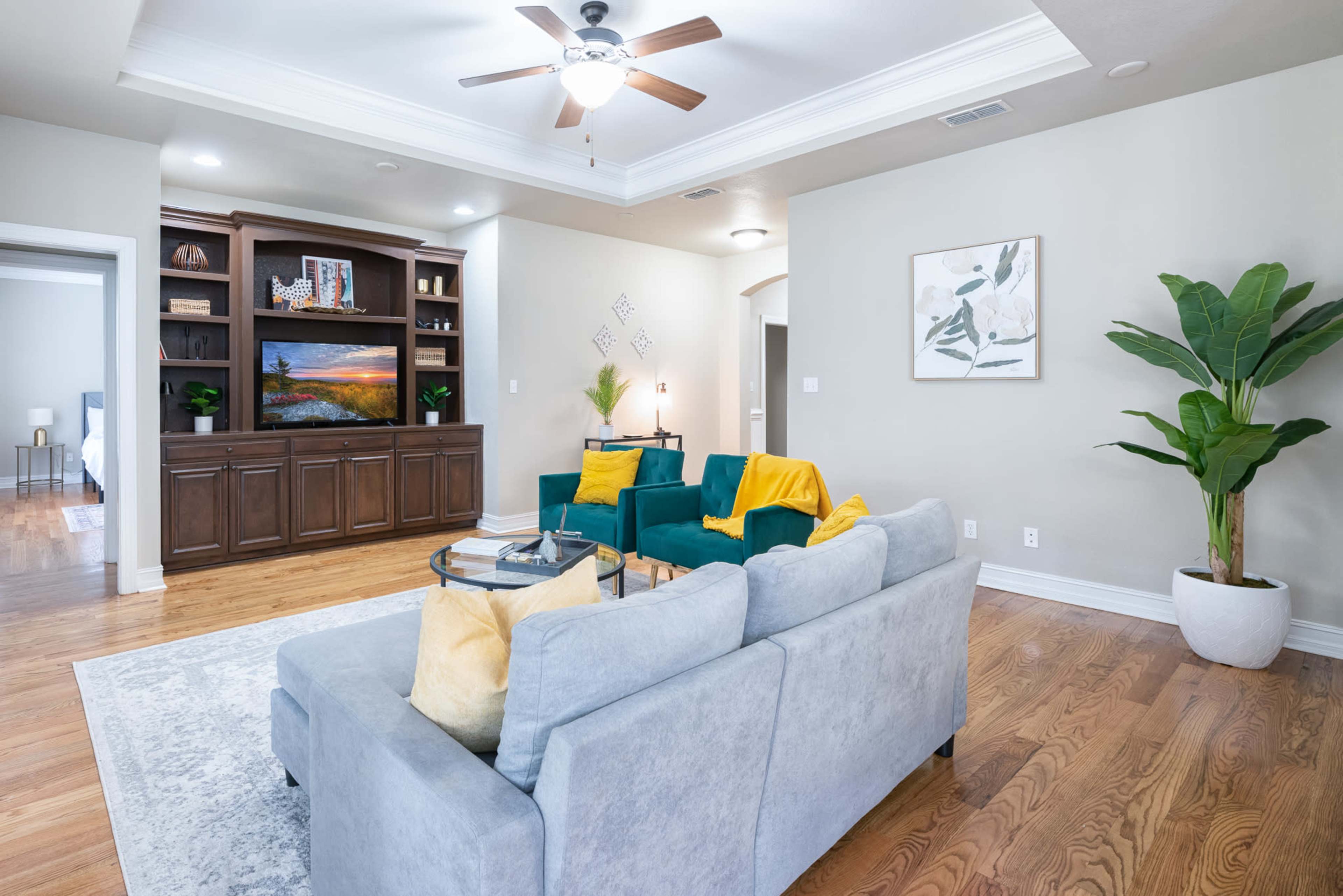 The living room features a light gray sofa, two teal chairs with yellow throws, a wooden entertainment center, and a large potted plant.