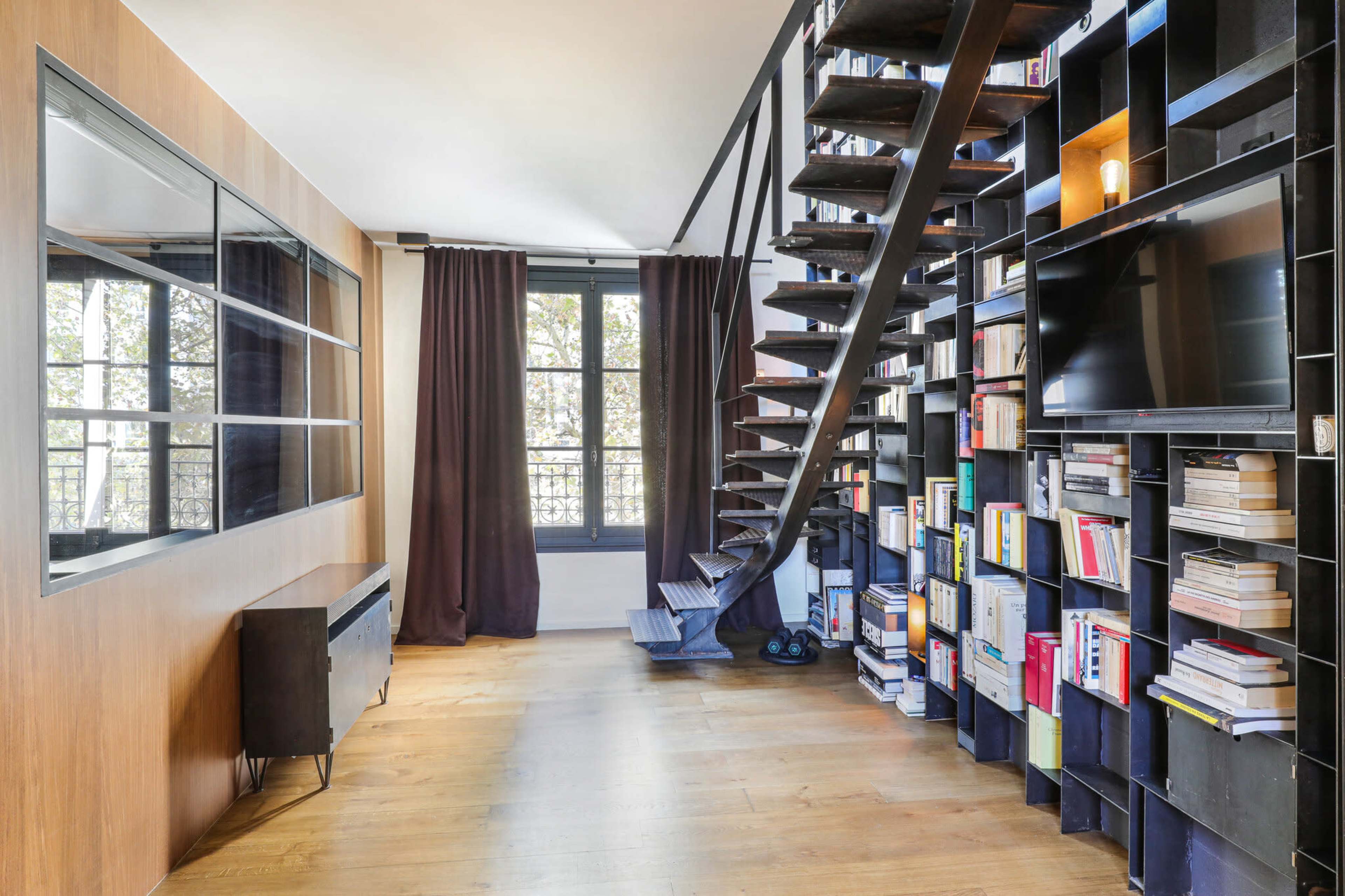 The image features a modern interior with a spiral staircase, a wall of bookshelves filled with books, and large windows allowing natural light to enter the space.
