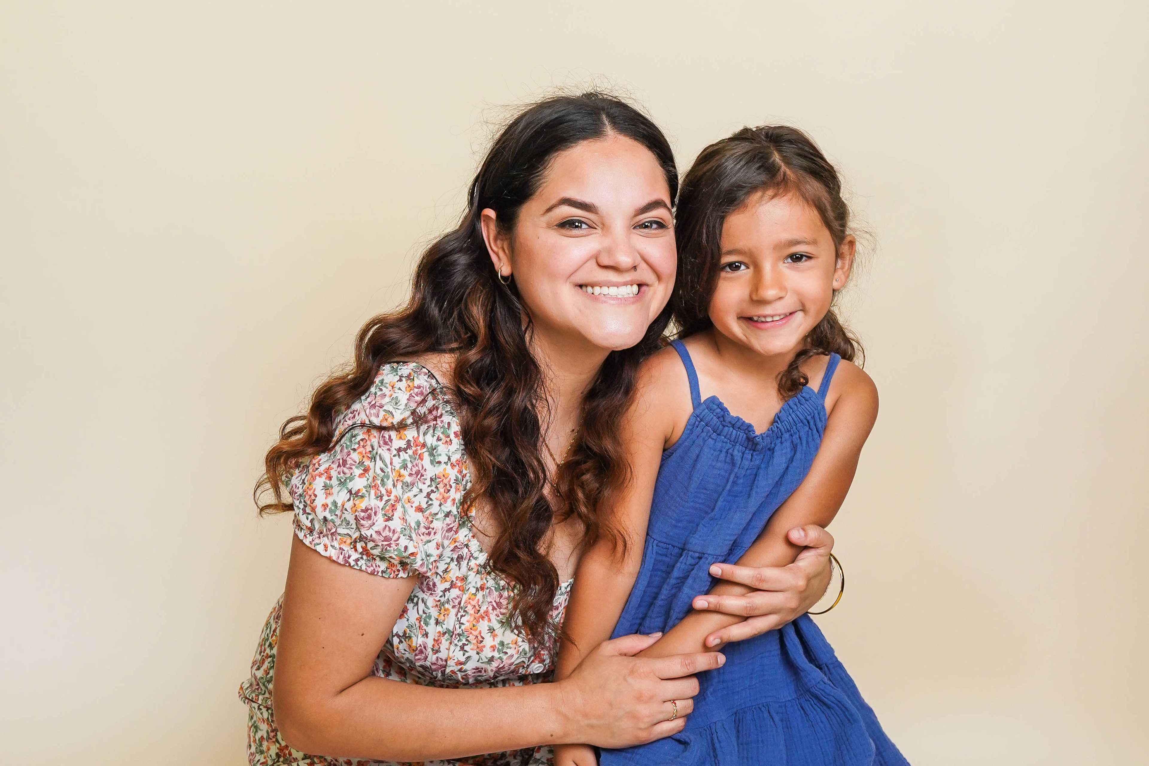 A woman and a young girl smile while sitting closely together in front of a light beige background.