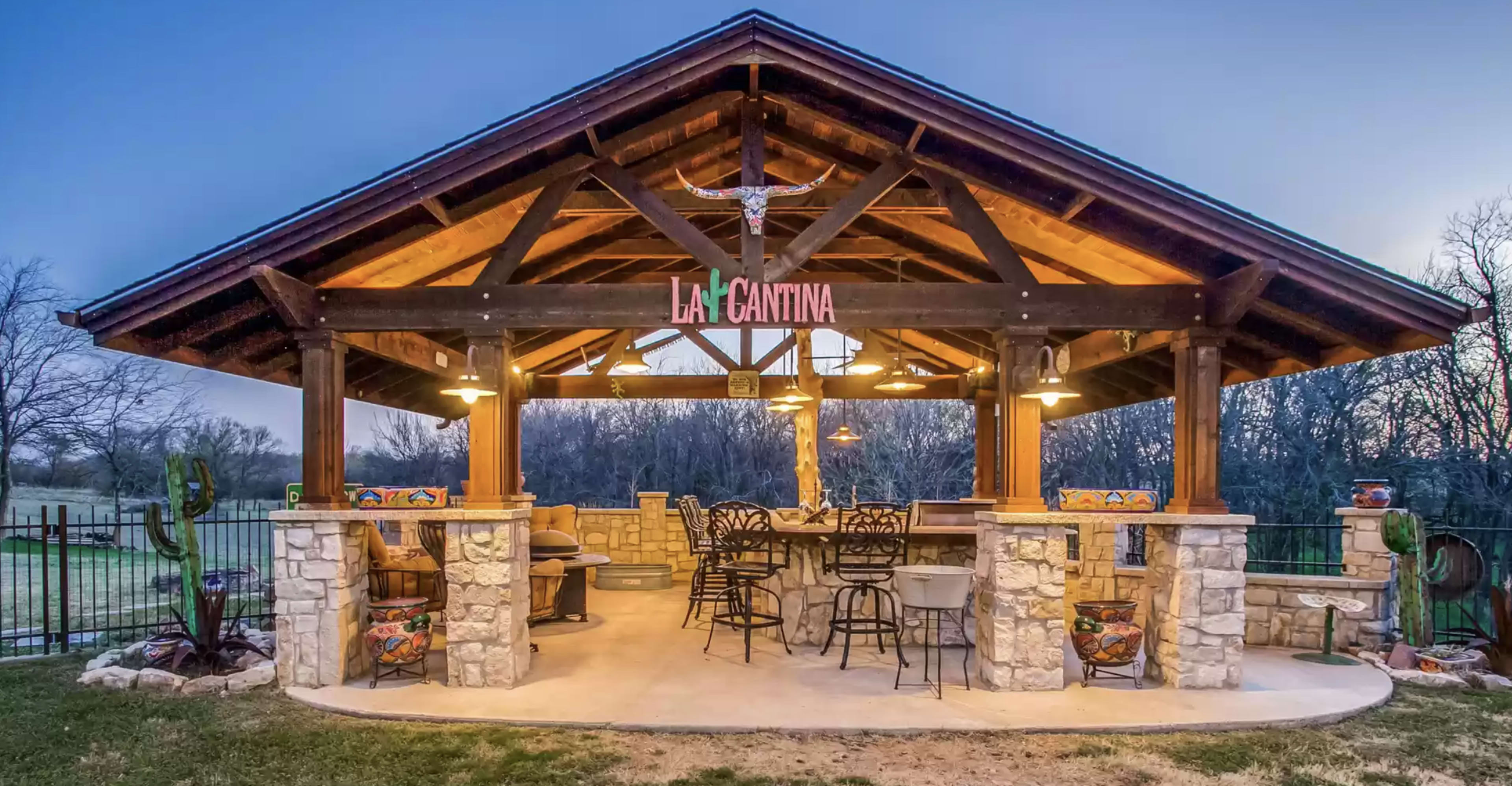 A rustic outdoor pavilion with a stone base, wooden beams, and bar seating under warm lighting.