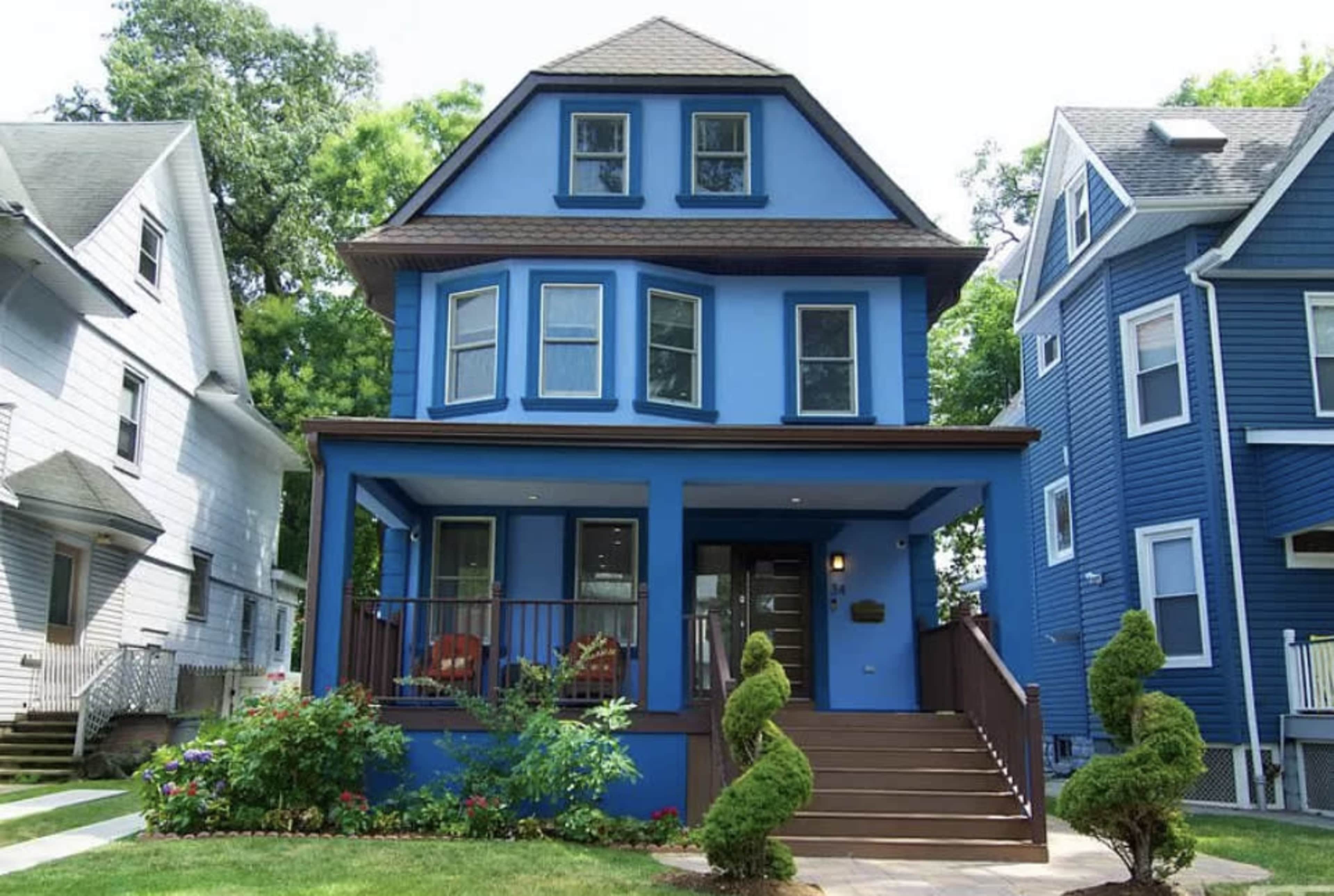 A blue three-story house with a front porch and landscaped steps is flanked by two other residential buildings.
