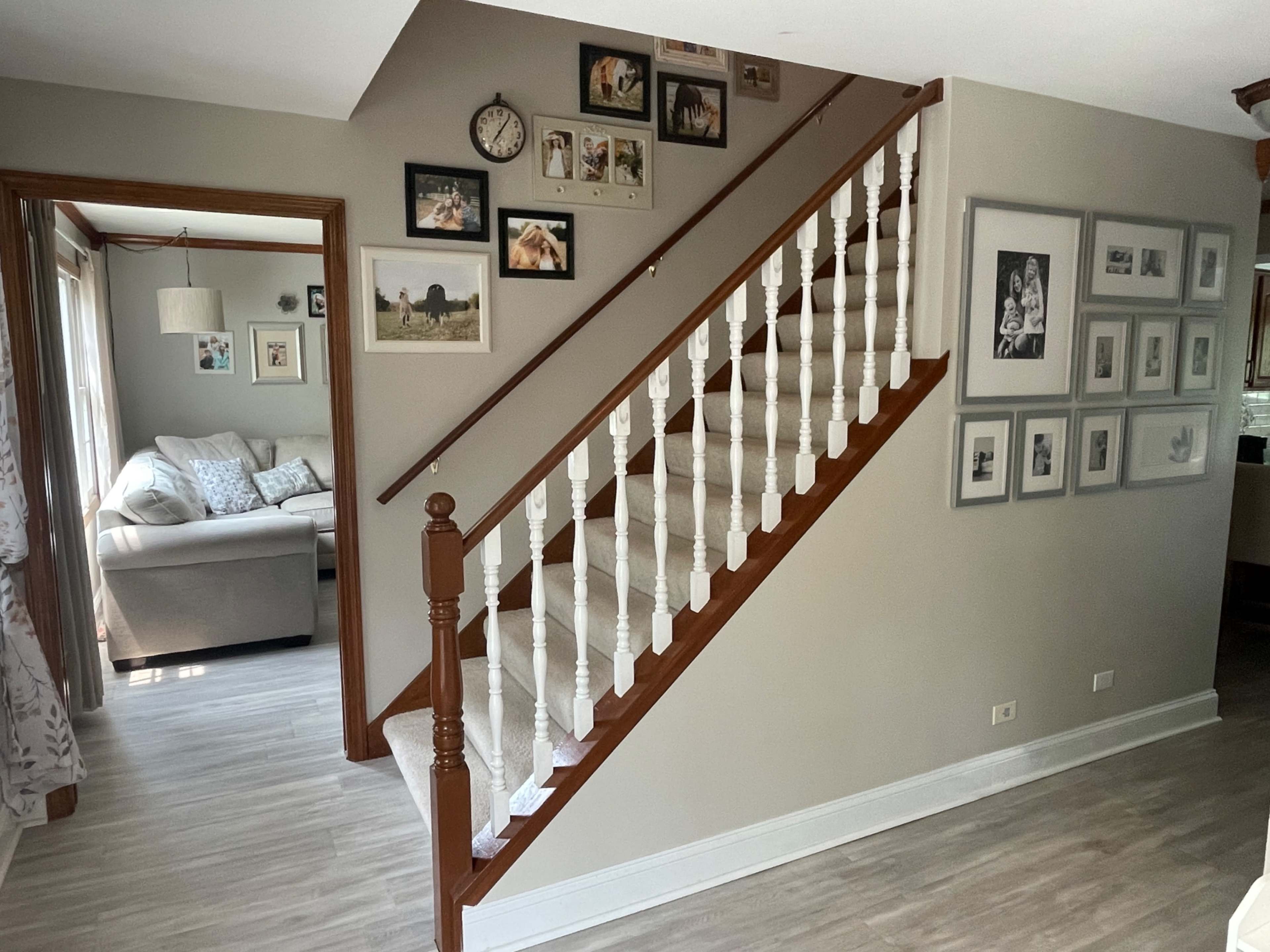 The image shows a staircase with white railings, leading to a second floor, adjacent to a living area with a sofa and framed photographs on the walls.